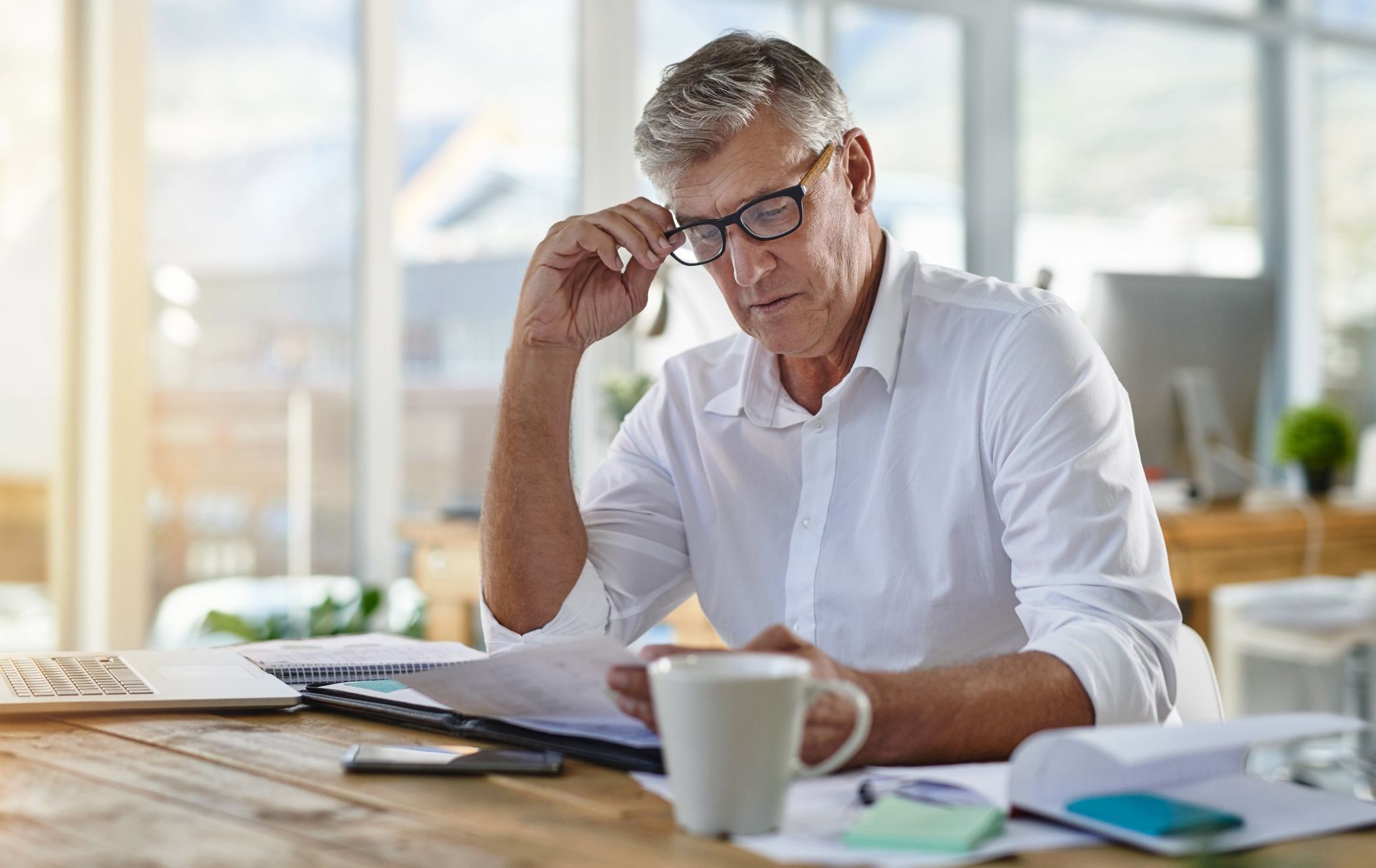 Person in a white shirt working at a desk with papers, laptop, and coffee mug by a bright window