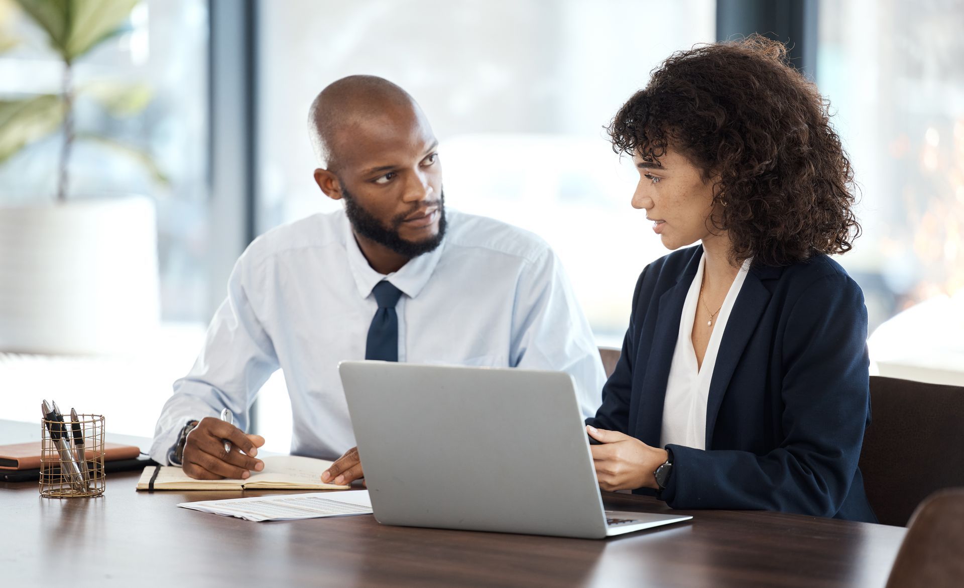 Two coworkers discussing over a laptop at a desk in a bright office
