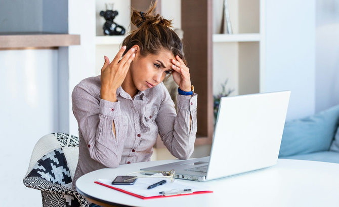 Person holding their temples while working at a laptop in a bright home office.