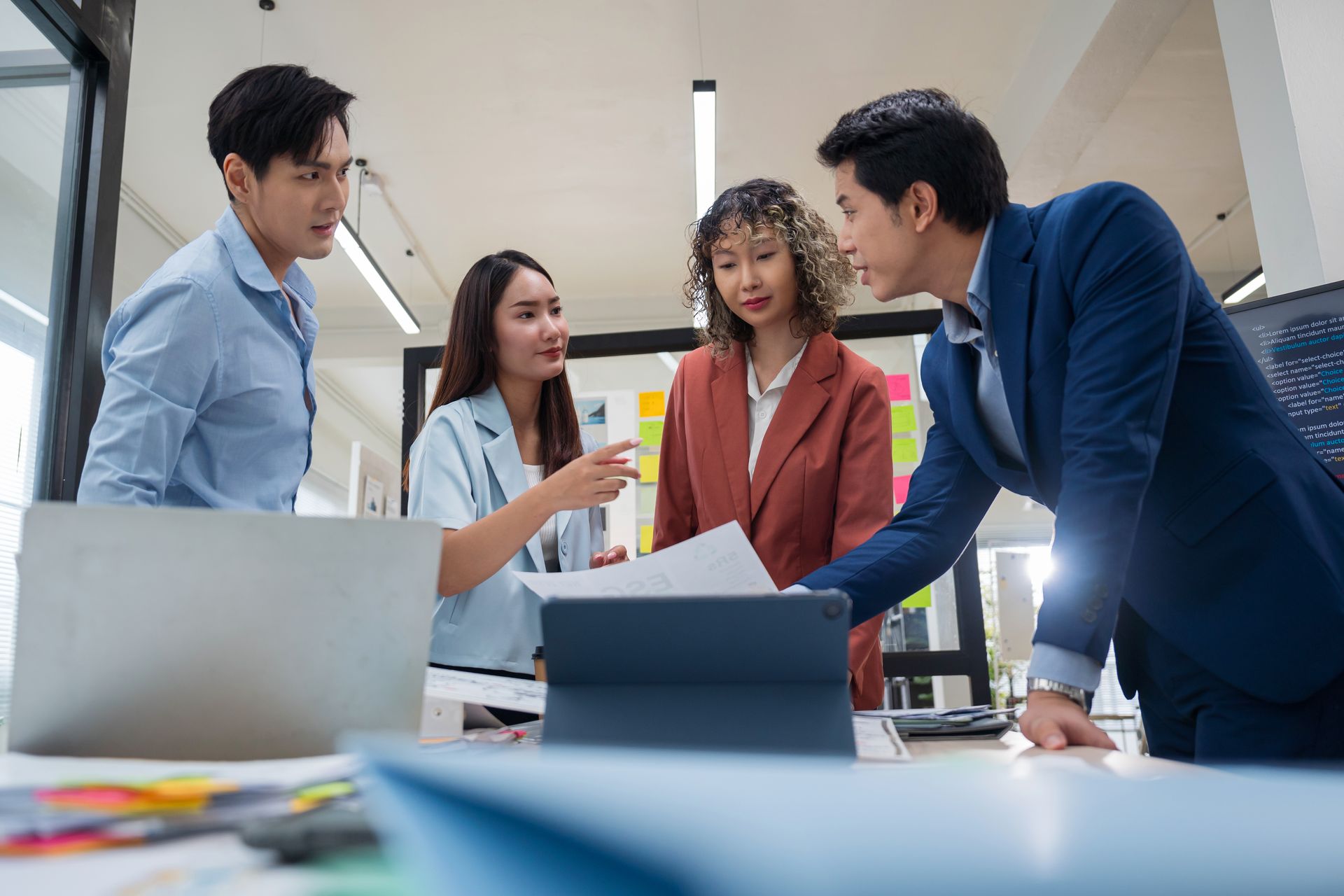 Four coworkers in a bright office lean over documents during a team discussion.