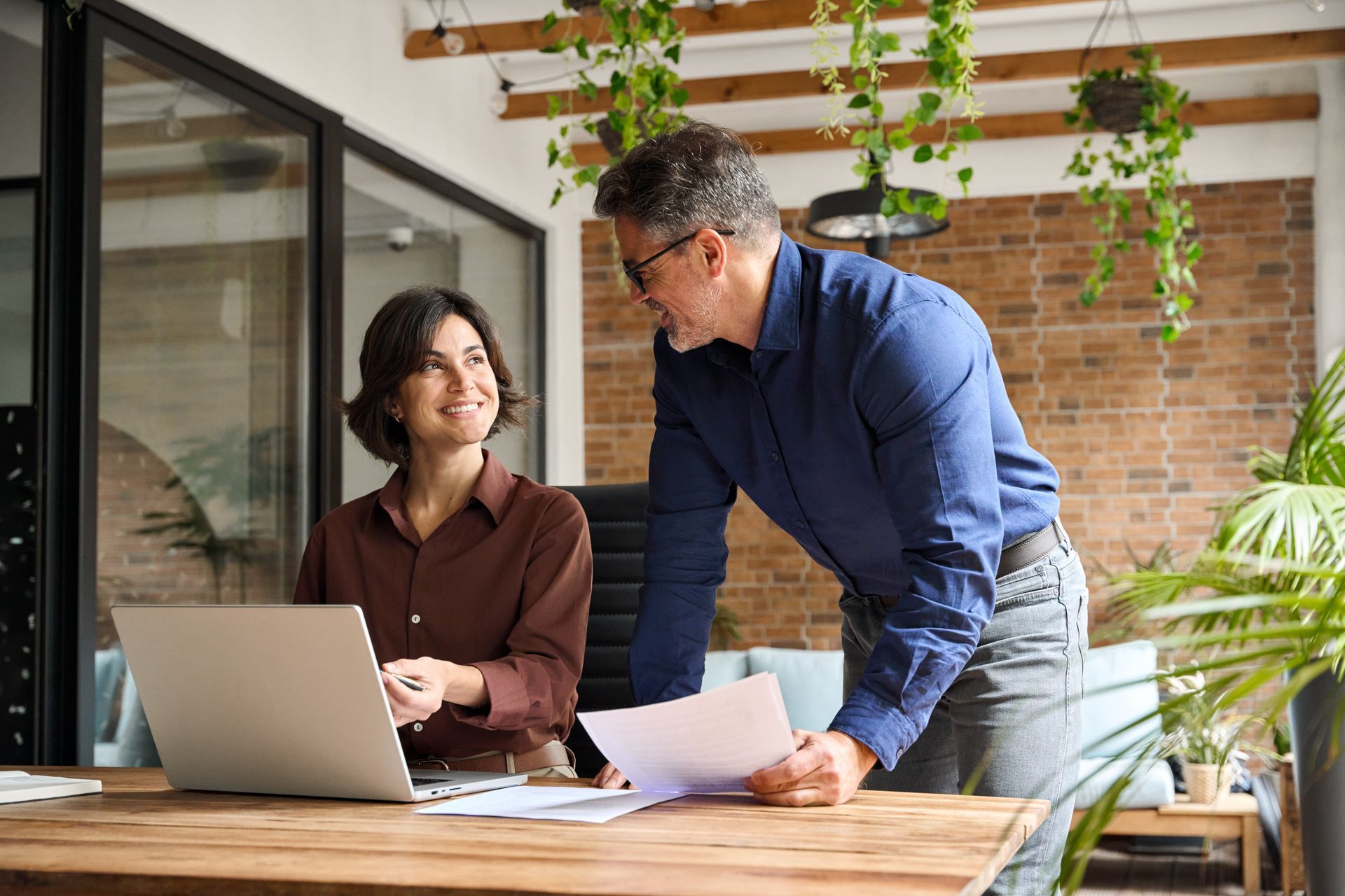 Two coworkers reviewing papers at a desk with a laptop in a bright office with plants