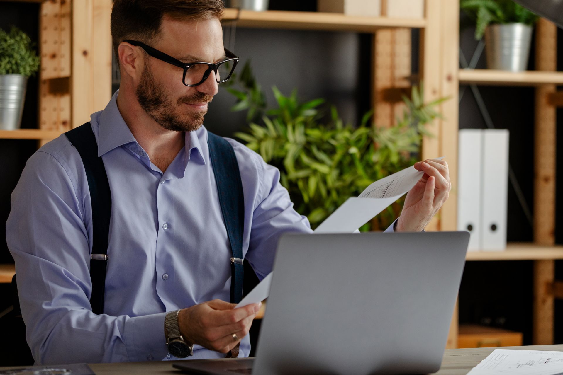 Man in glasses reviewing a document at a laptop in an office with shelves and plants.