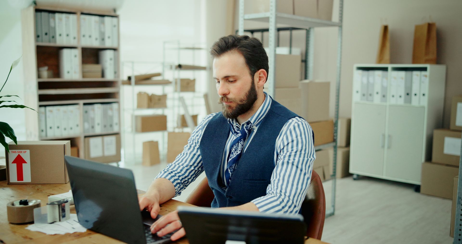 Man working on a laptop at a desk in a bright office with shelves, boxes, and filing cabinets.