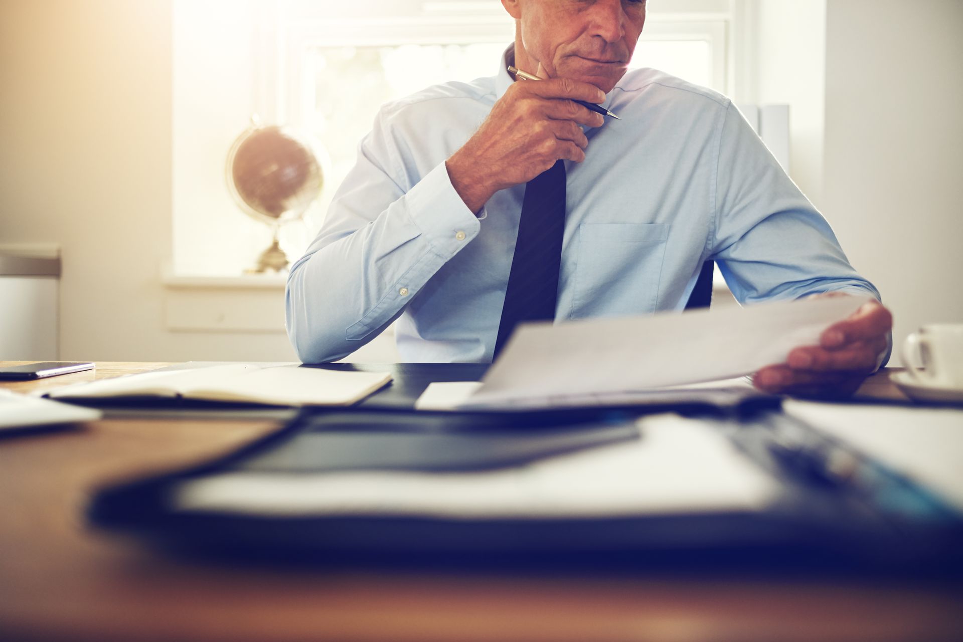 Man in a light blue shirt reviewing paperwork at a desk, hand on tie, in a bright office