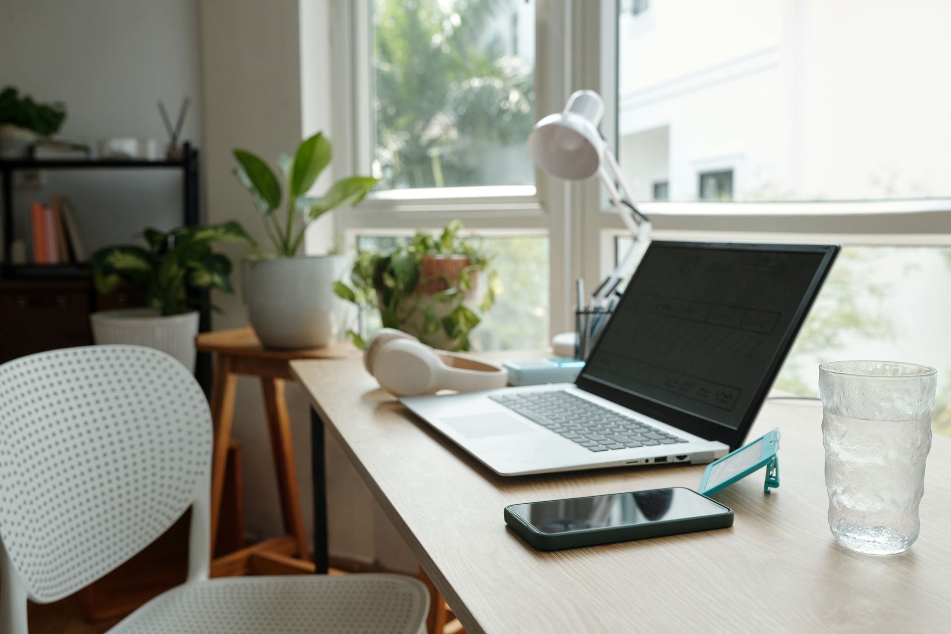 Home office desk with open laptop, desk lamp, plants, notebook, phone, and glass of water by a bright window