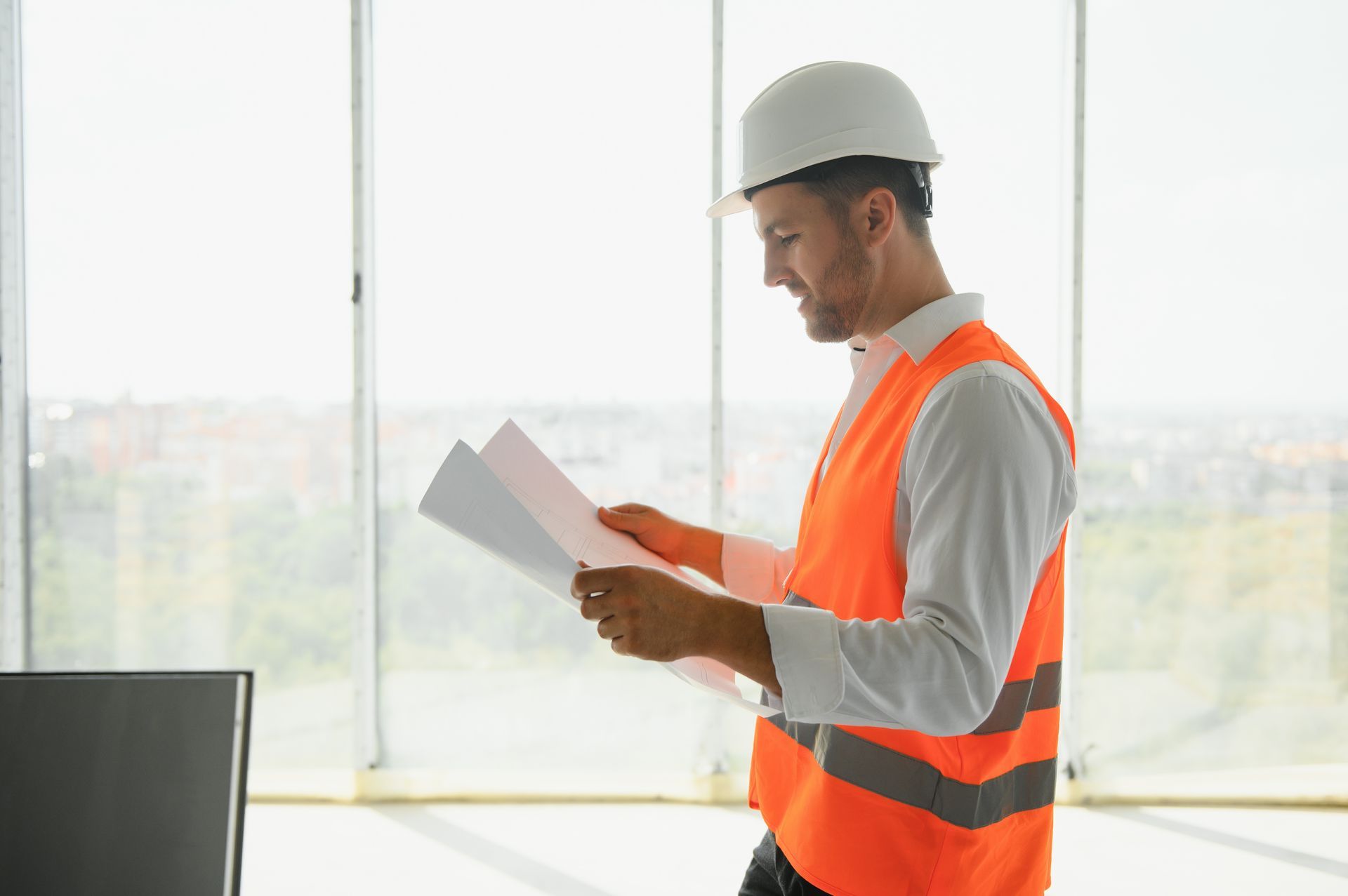 Construction worker in orange vest and hard hat reviewing papers by a window