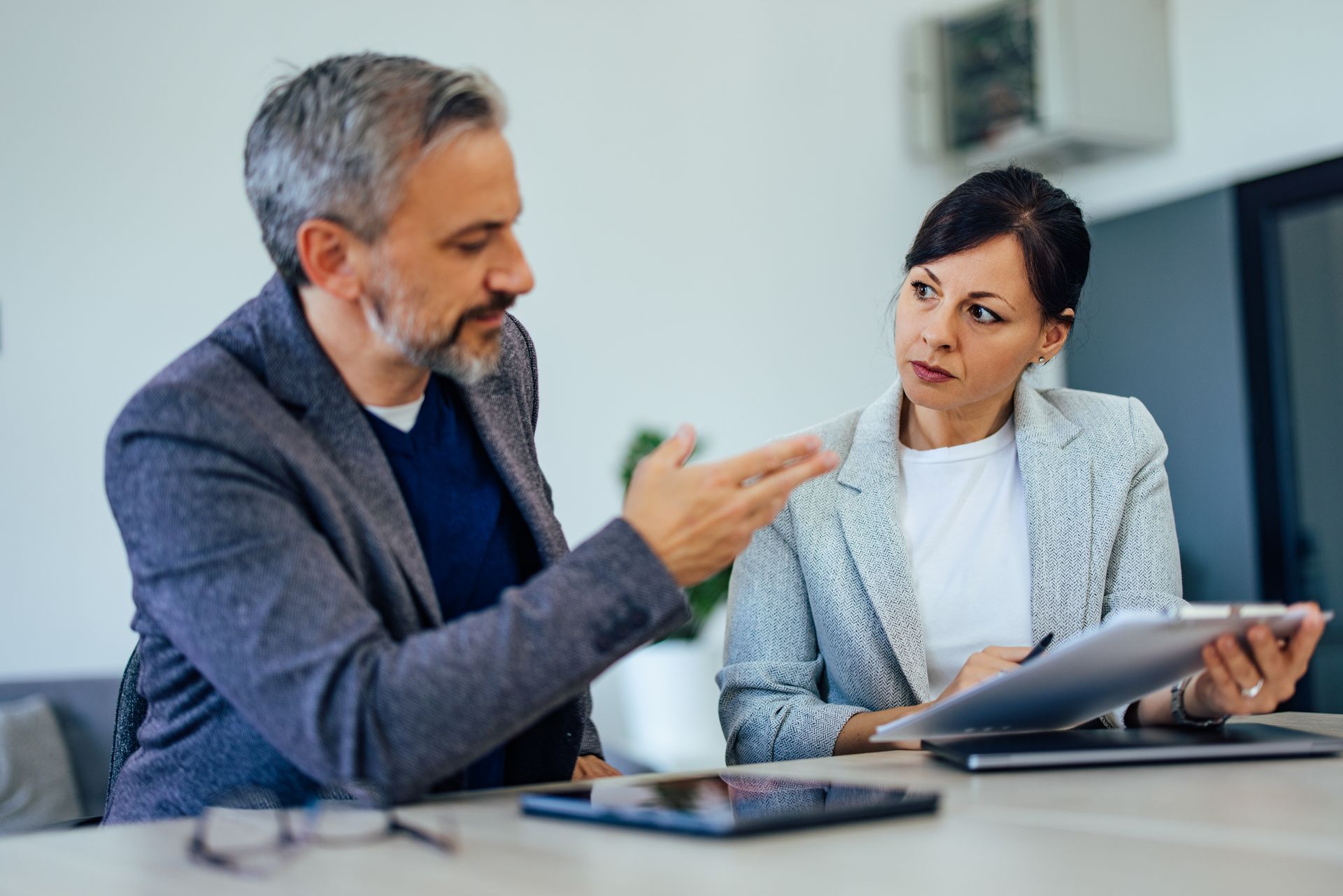 Two coworkers in a meeting, one gesturing while the other reviews documents at a table.