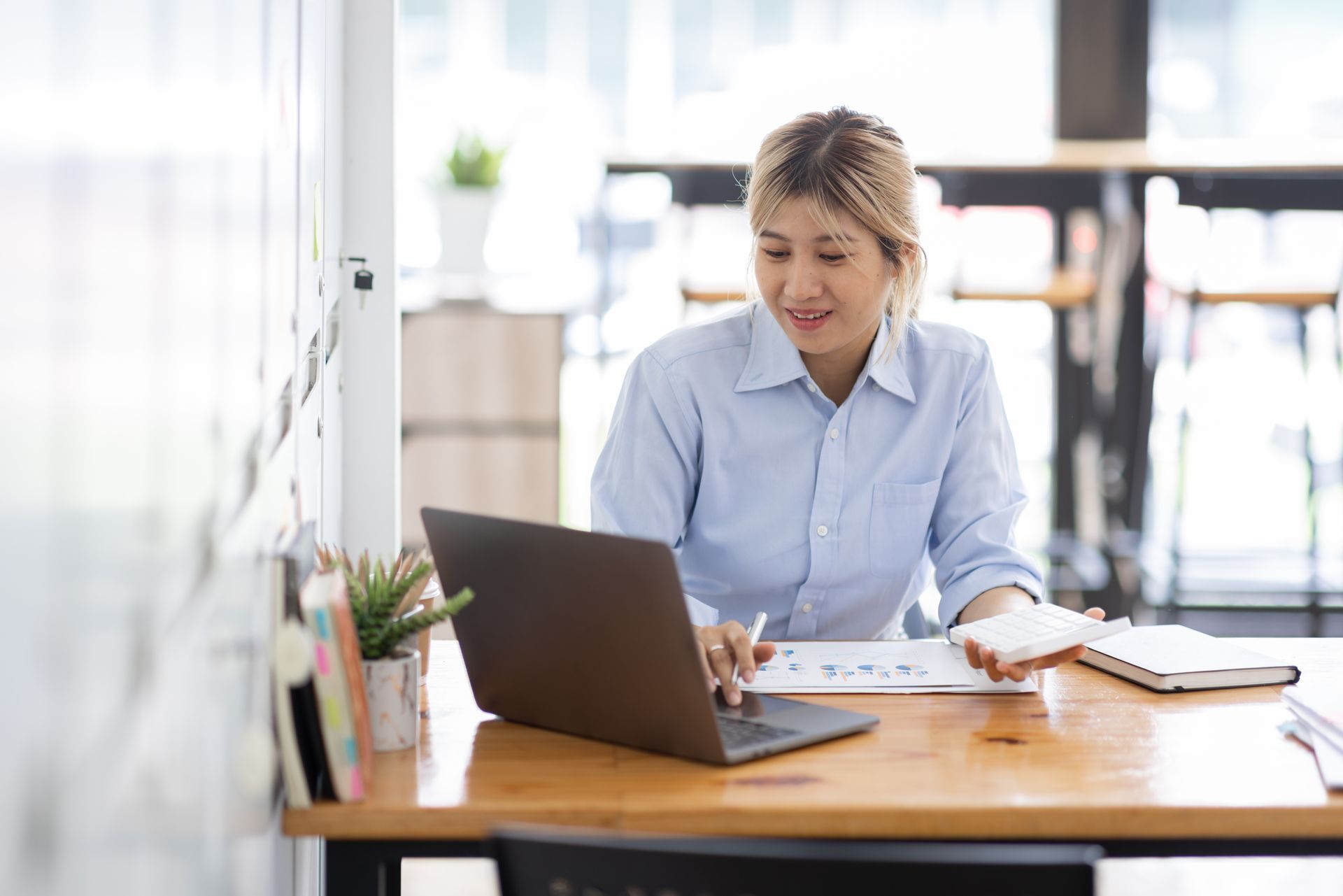 Woman working on a laptop at a desk in a bright office, with papers and a notebook nearby.