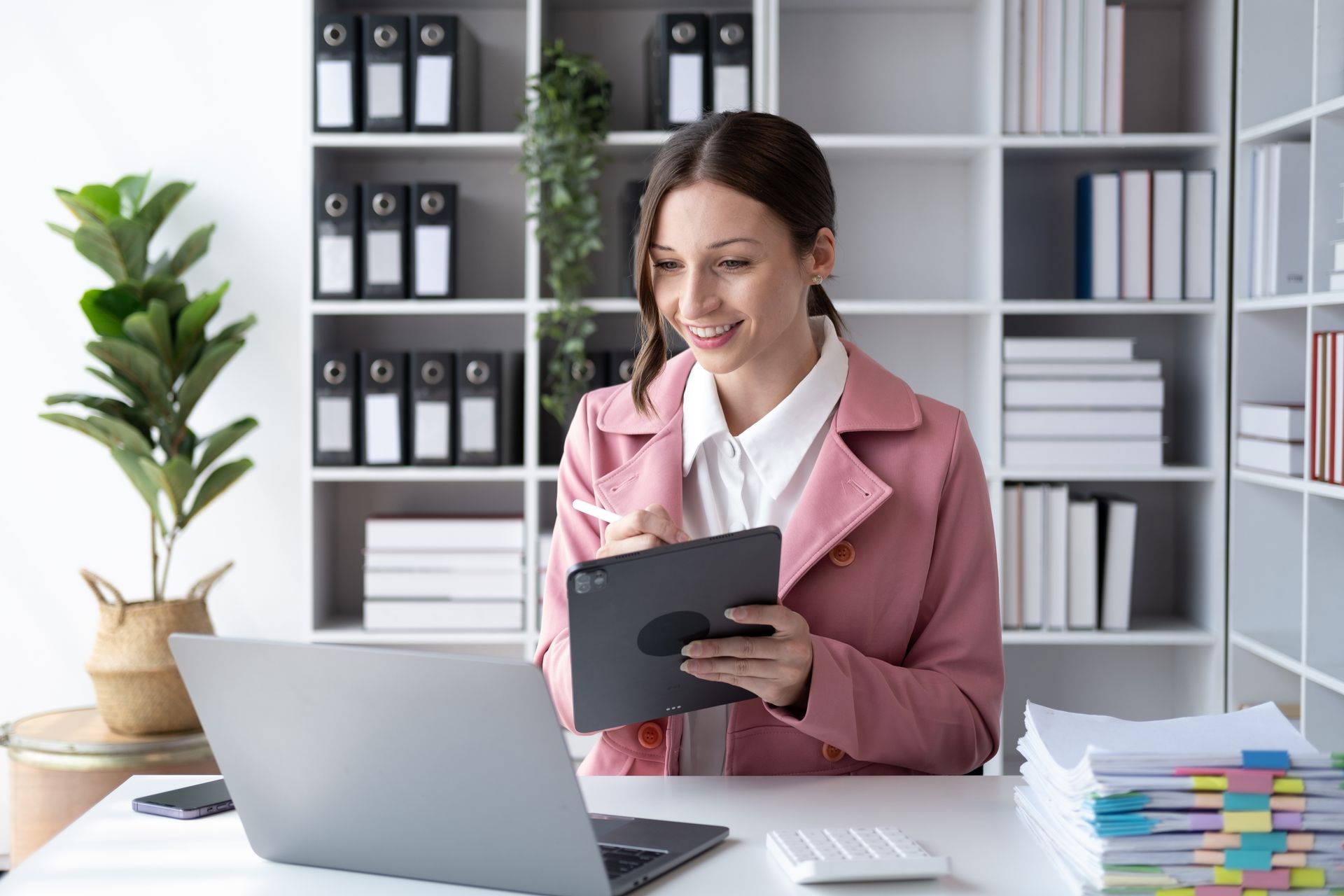 Woman in a pink blazer reviewing notes beside a laptop in a tidy office with shelves and binders.