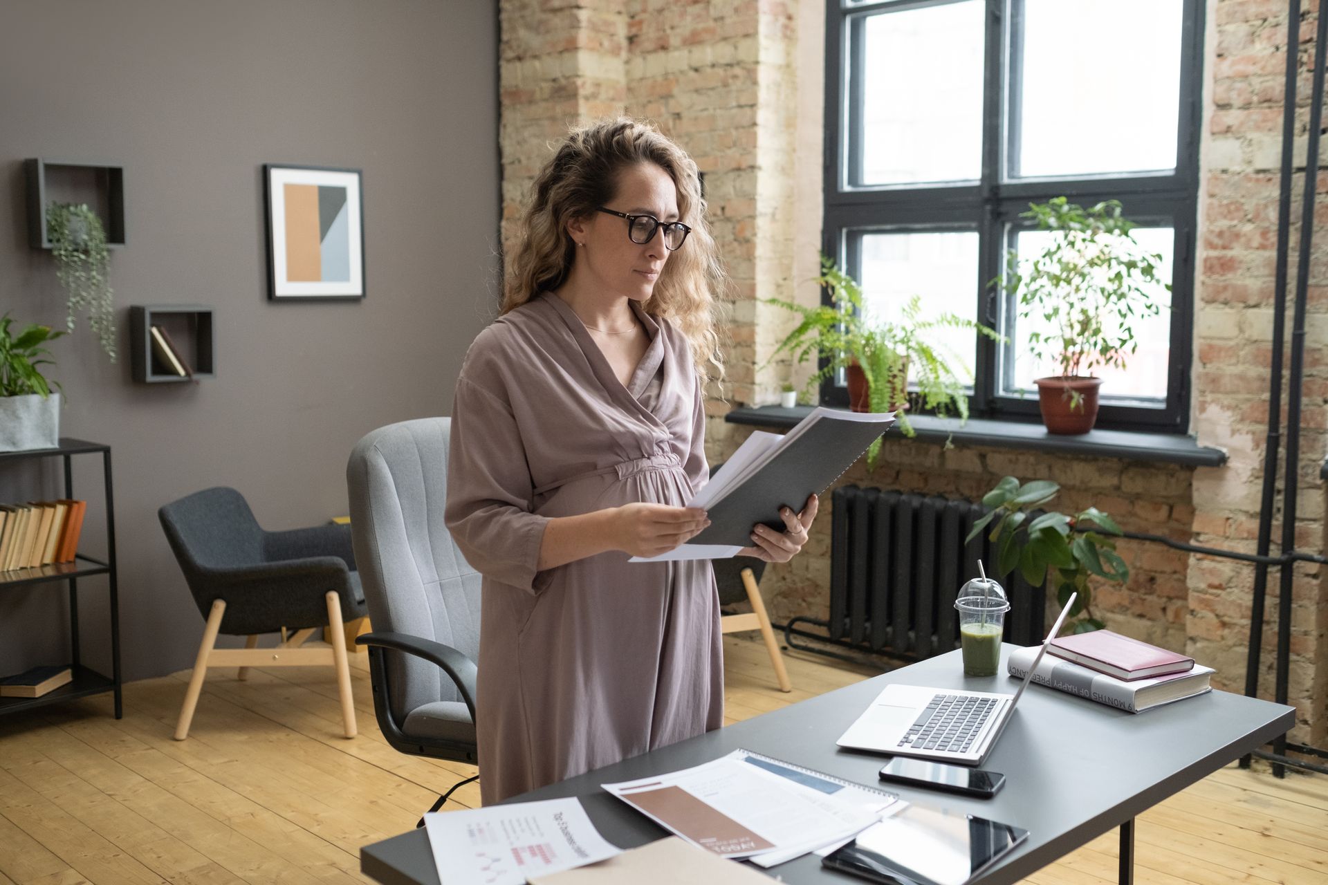 Woman standing at a desk in a bright office, holding a tablet near a laptop and papers by a window.