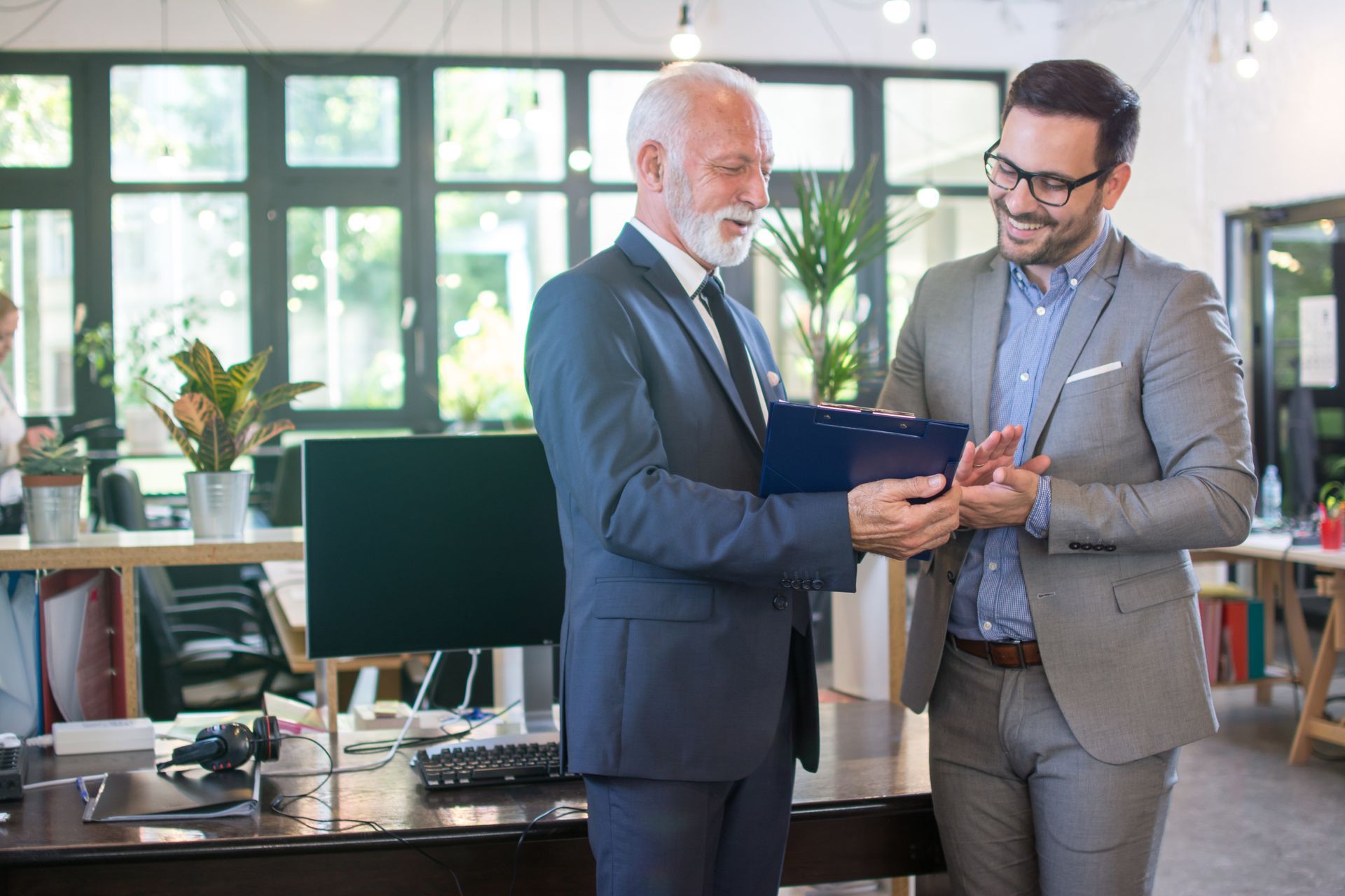 Two professionals in business attire stand in an office, reviewing notes on a clipboard and discussing a project.