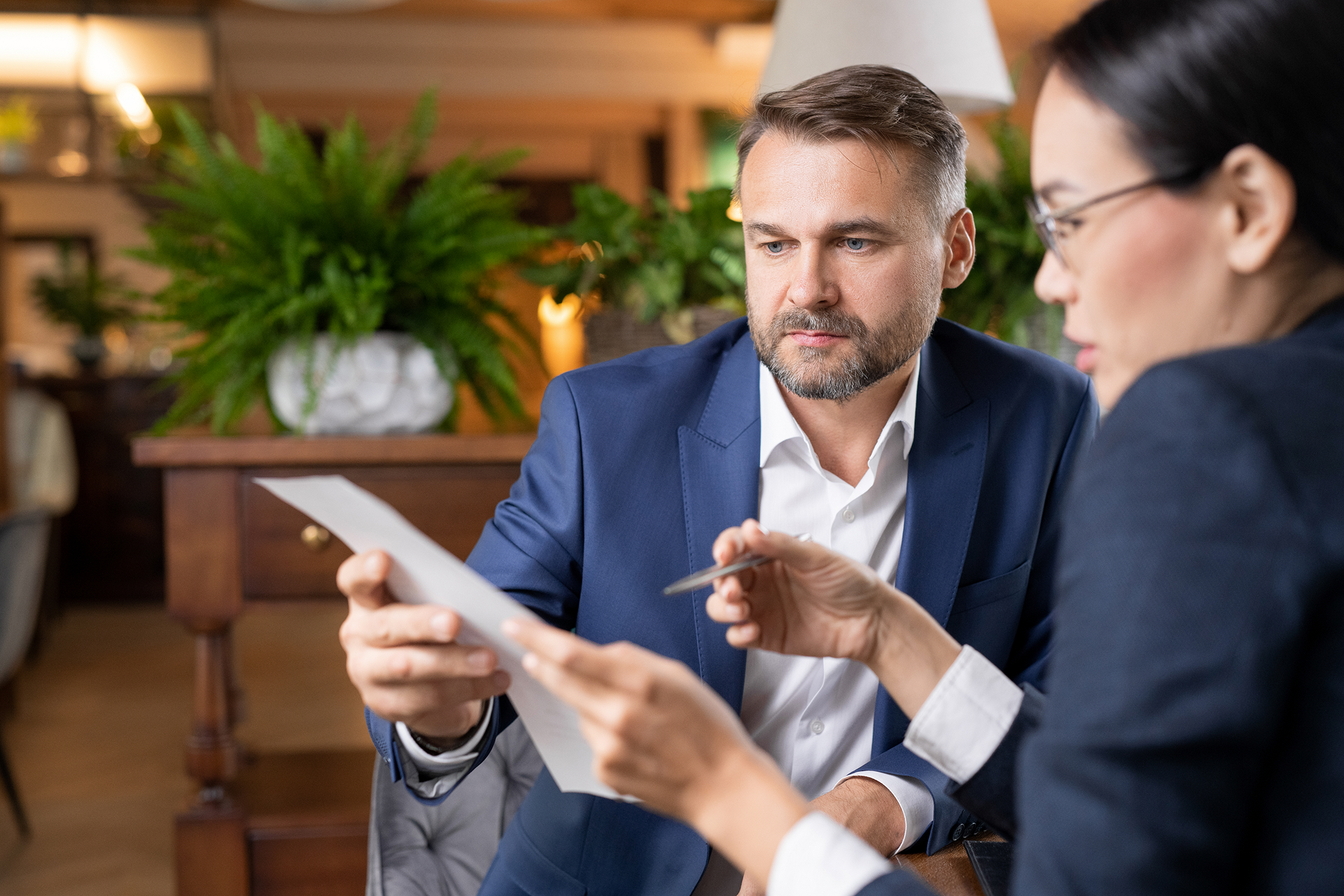 Two business professionals in blazers review a document together in a brightly lit, plant-filled office setting.