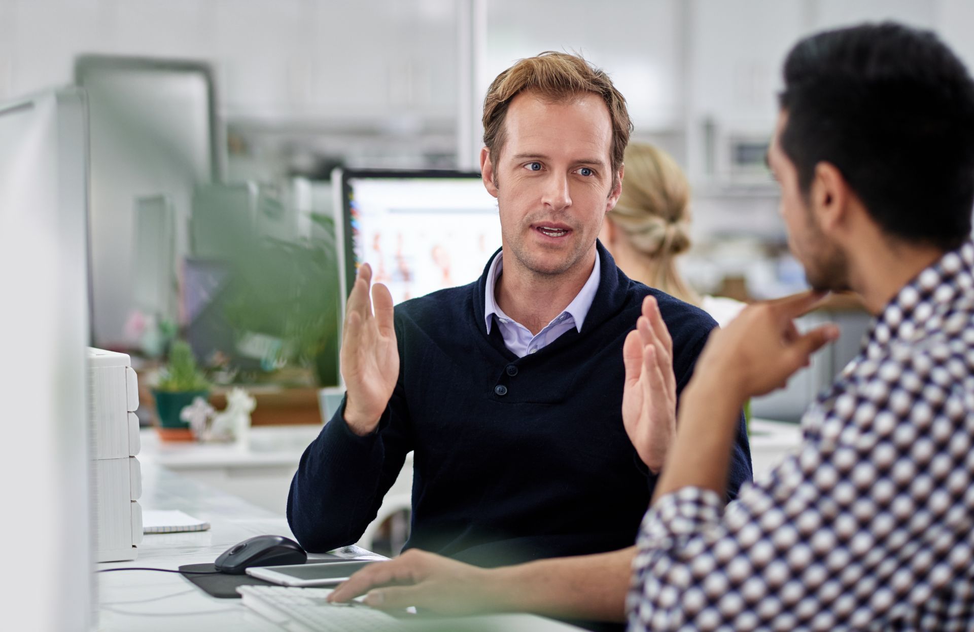 Two people talking in an office, one gesturing while seated at a desk with a computer monitor nearby