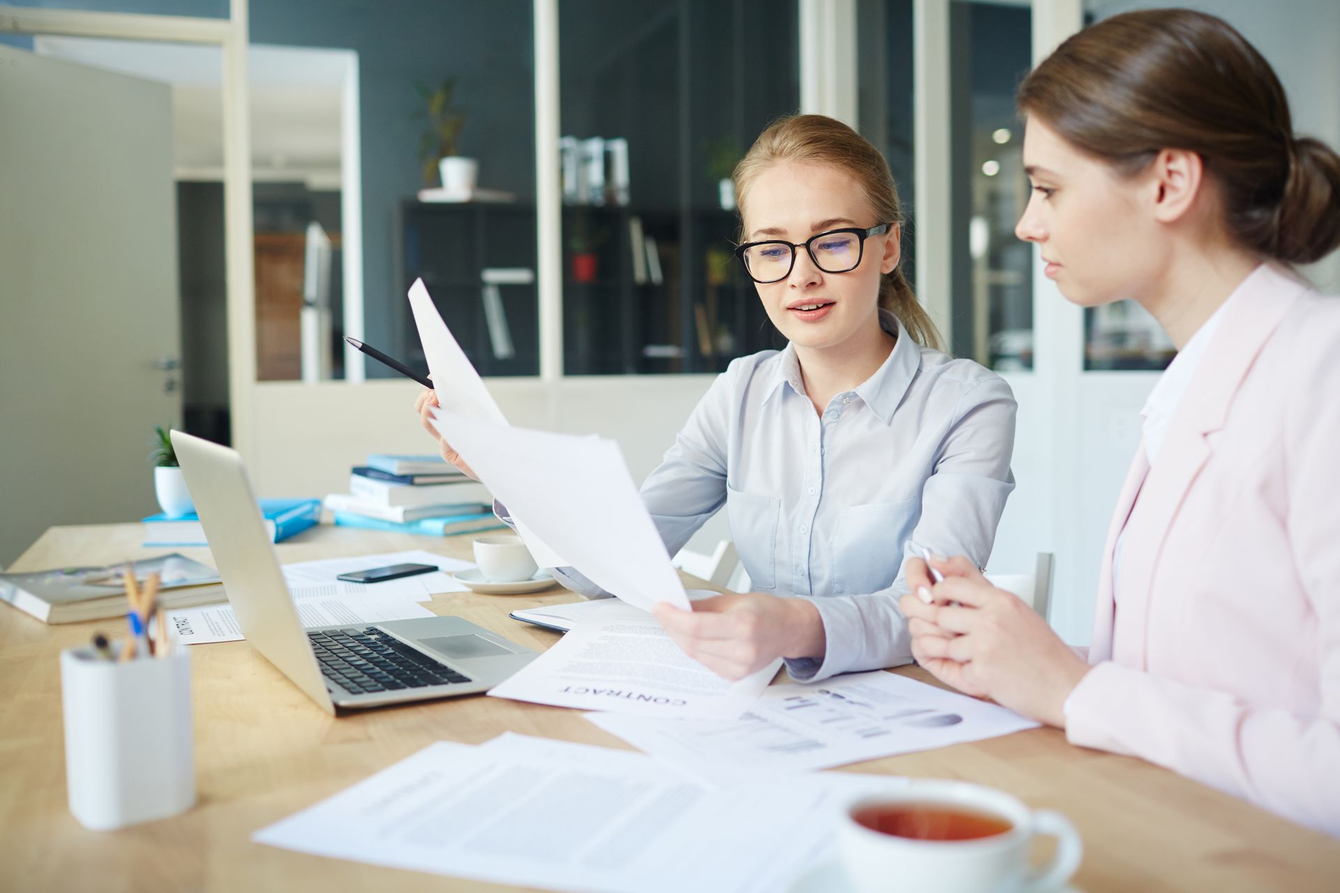 Two coworkers discussing documents at a desk with laptops in a bright office