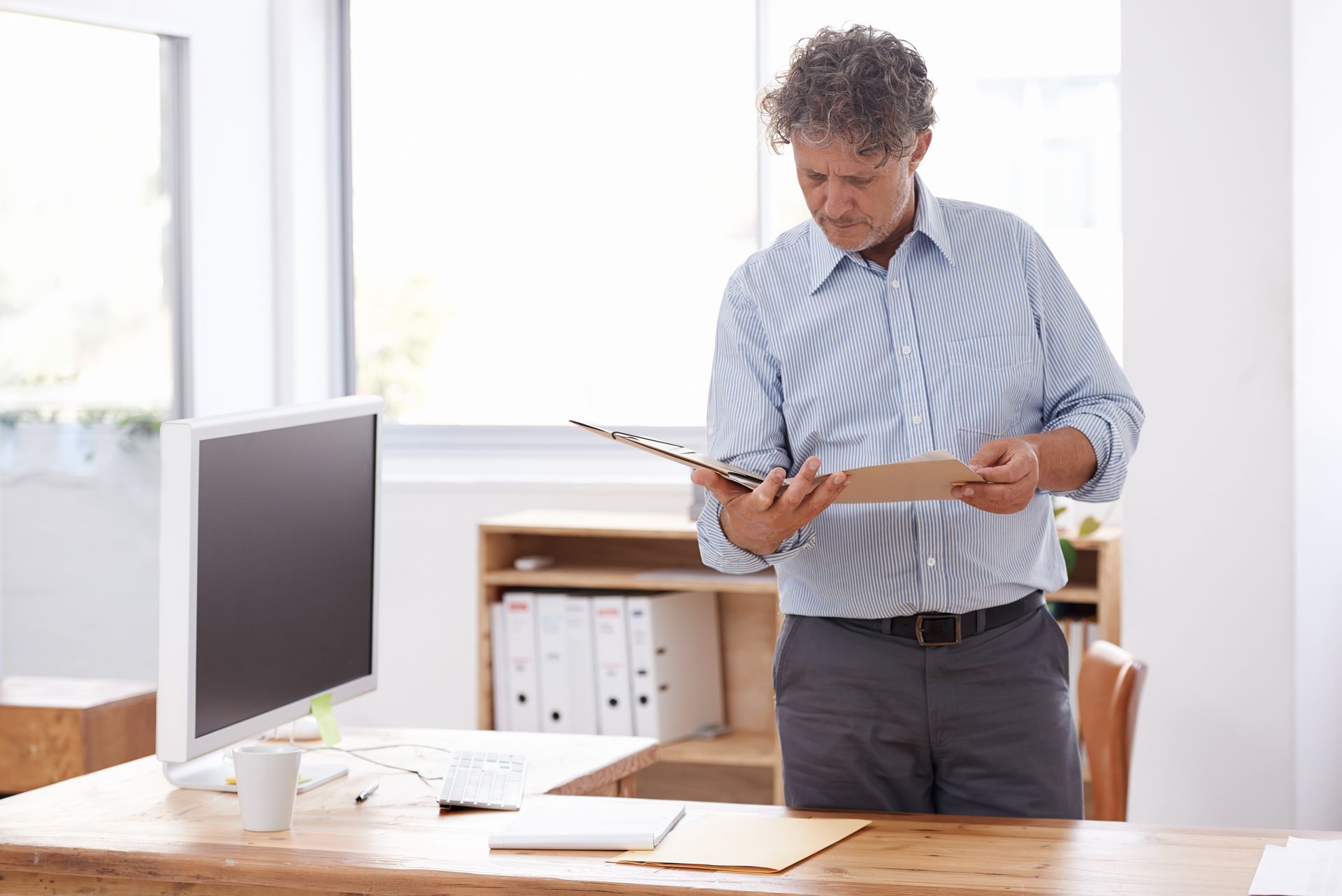 Man examining papers at a desk in a bright office, with a computer monitor nearby.