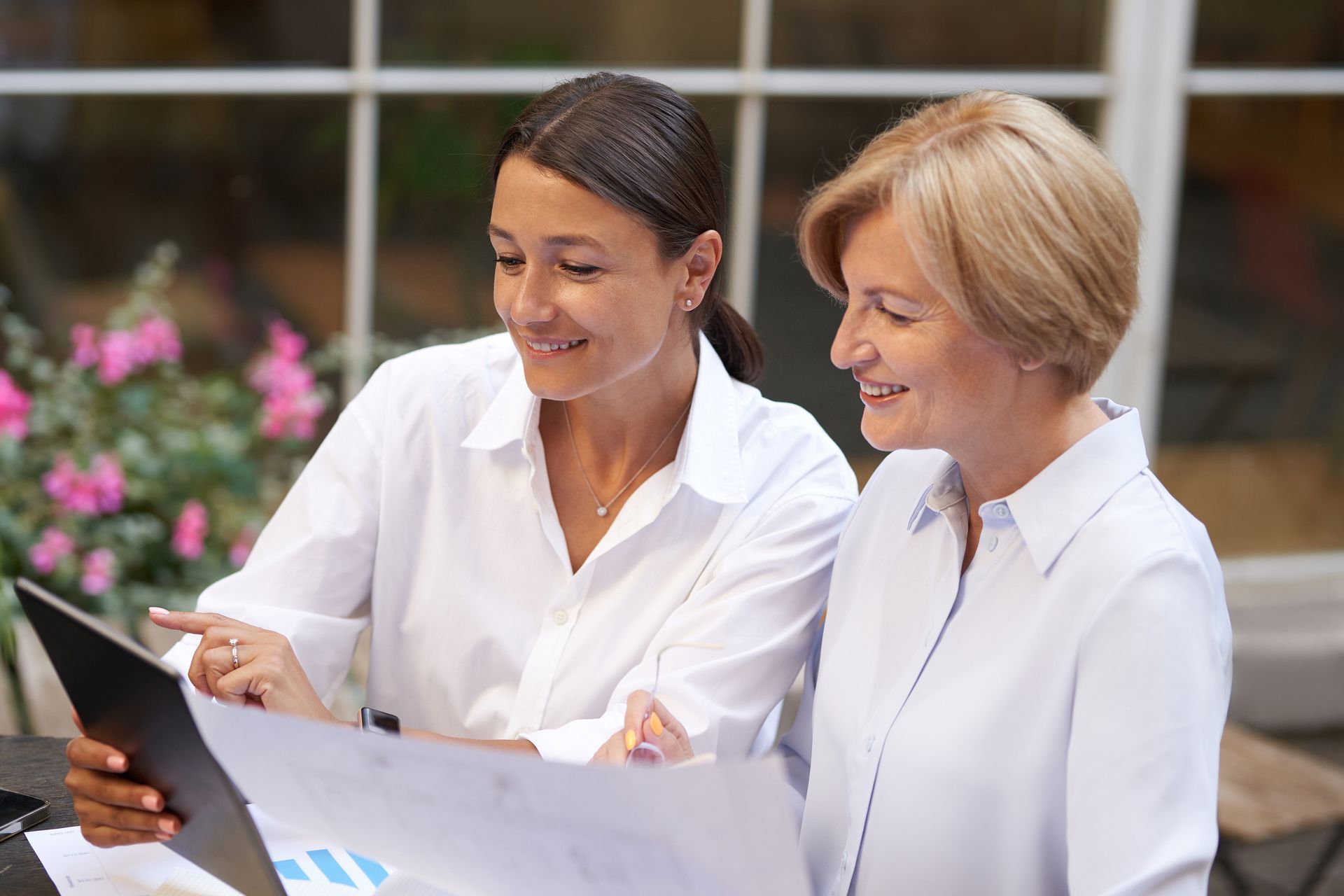 Two women in white shirts reviewing documents together at a table with a laptop.