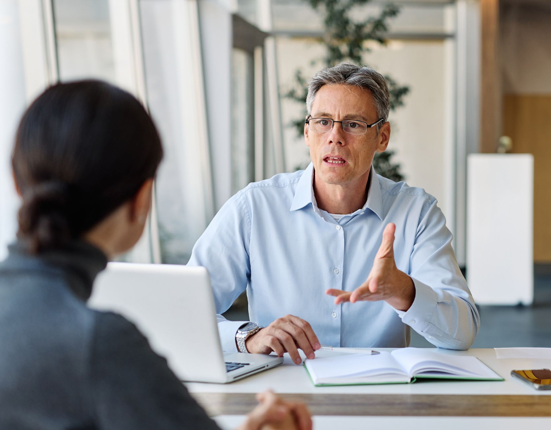 Two people in a bright office meeting, one speaking and gesturing across a laptop and notebook.