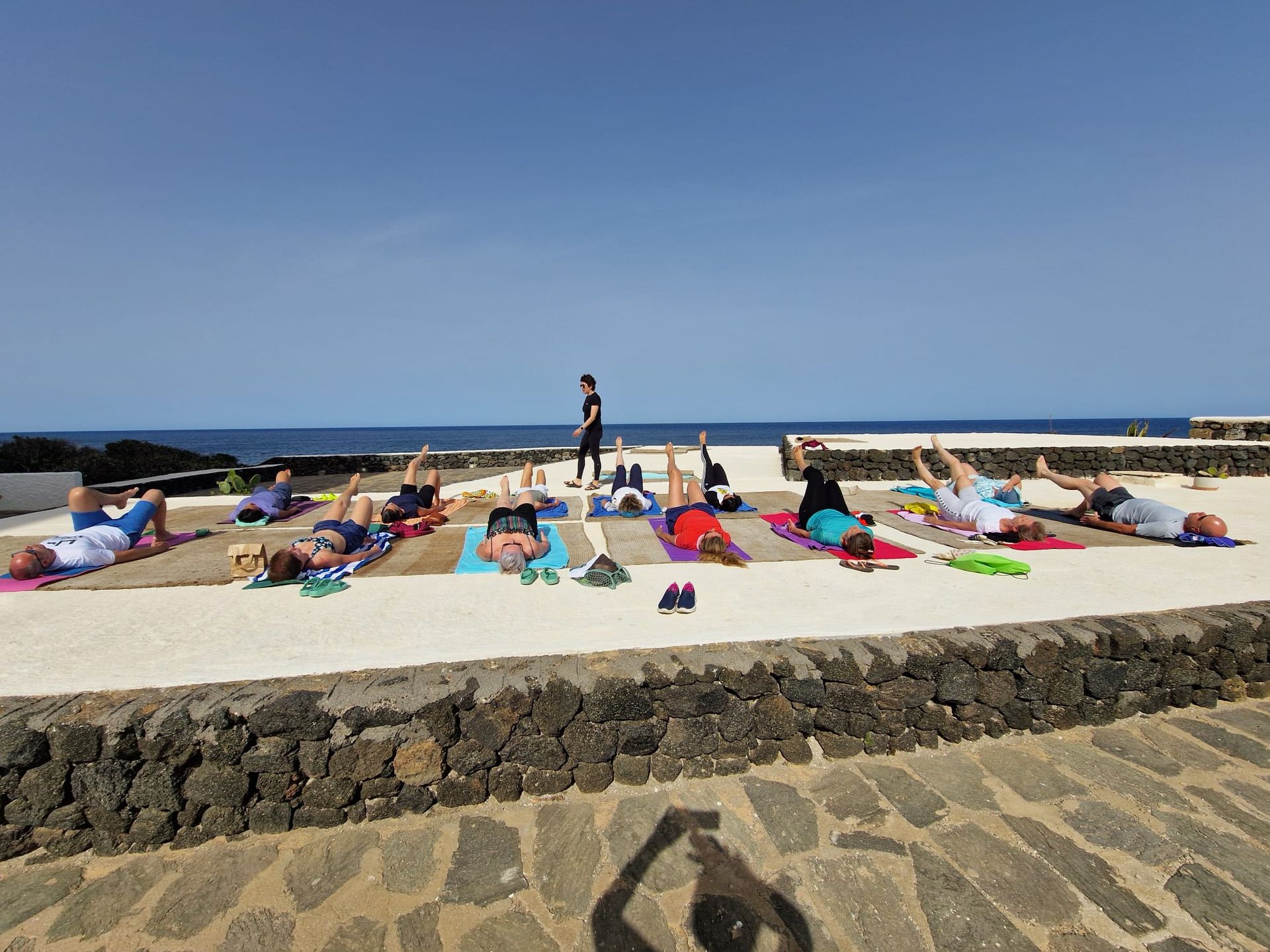 Persone che fanno yoga all'aperto su tappetini, l'istruttore al centro, sullo sfondo l'oceano, giornata di sole.