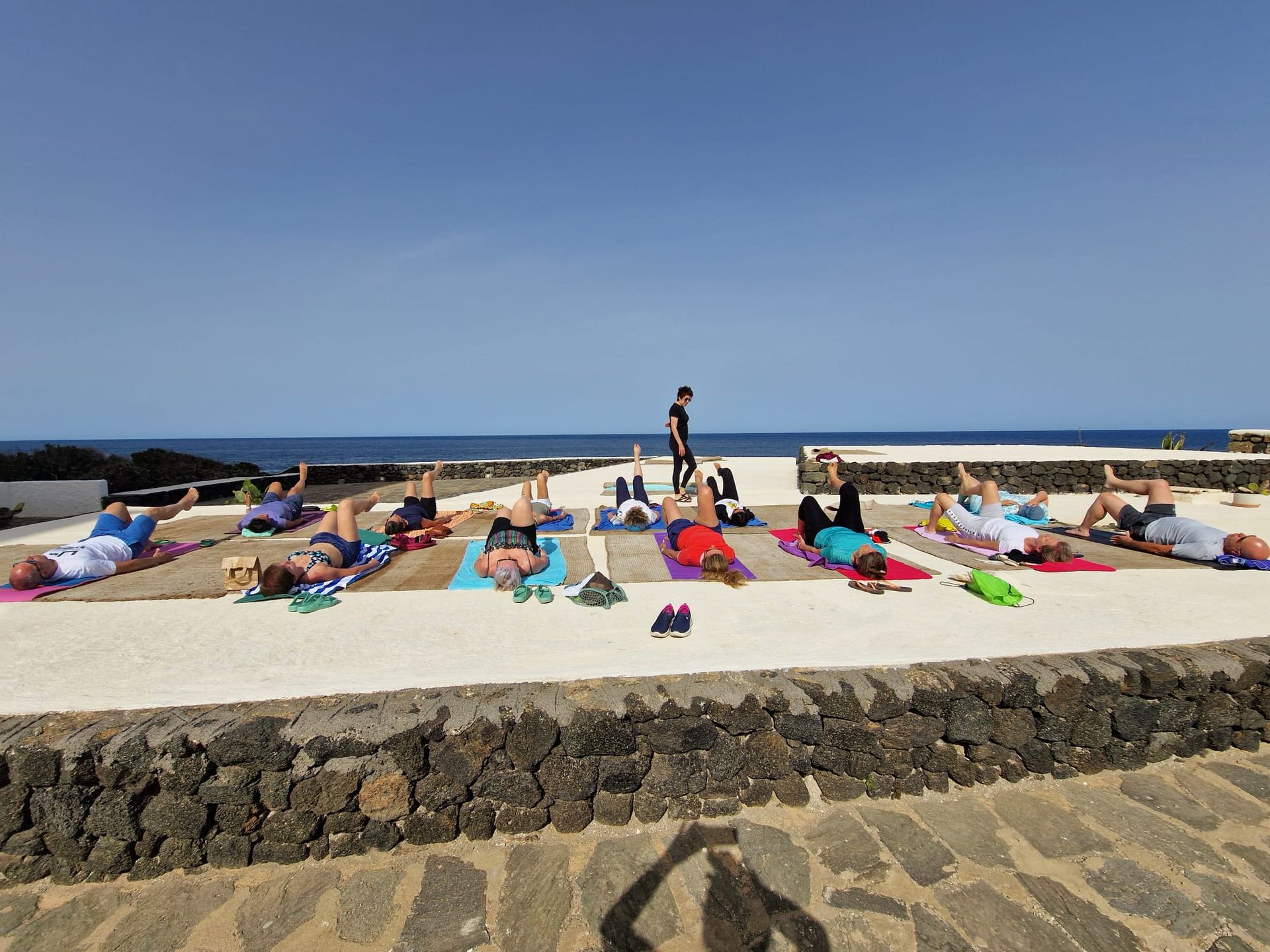 Persone che fanno yoga su tappetini all'aperto, con l'oceano sullo sfondo e un cielo azzurro.