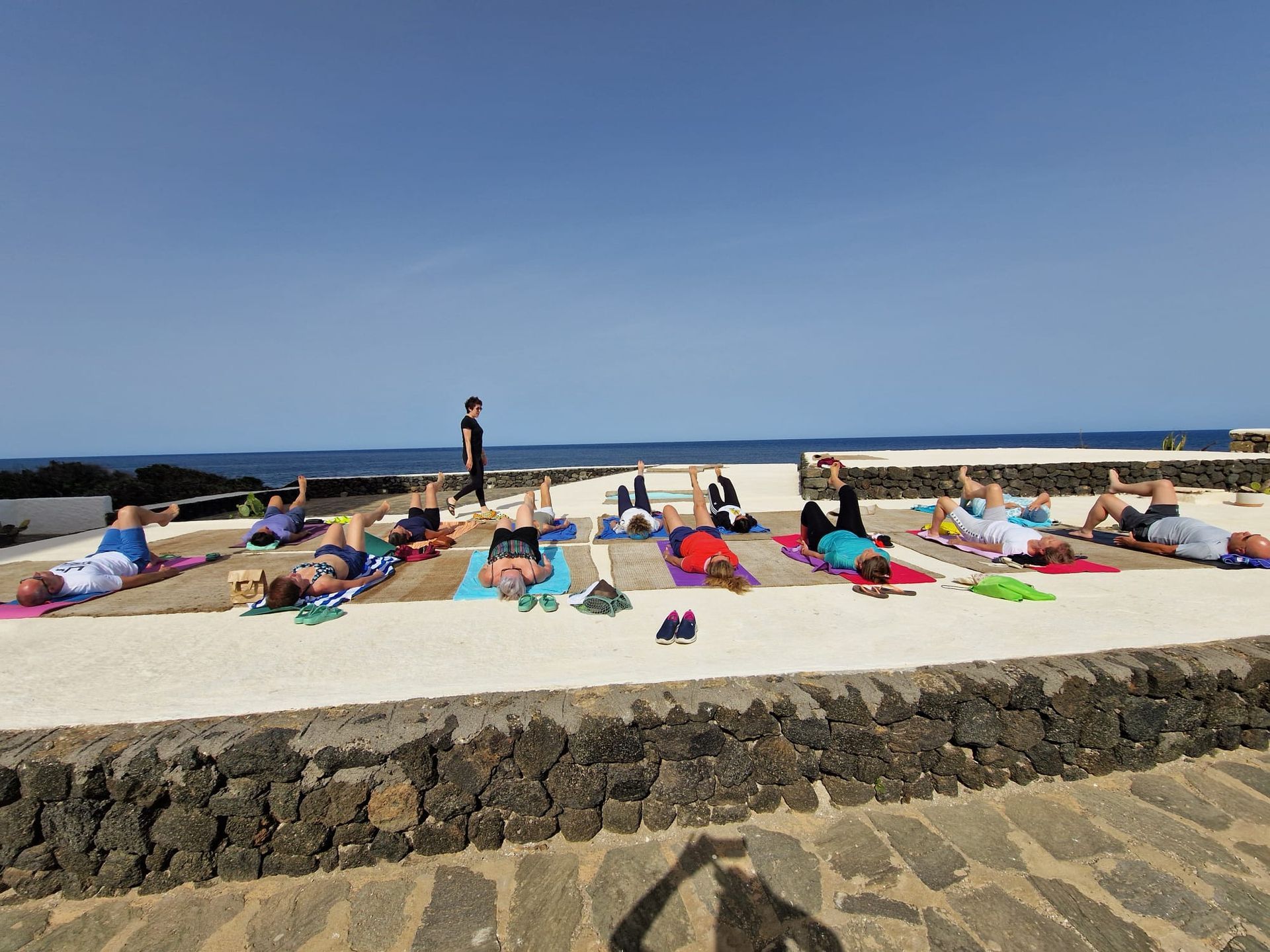 Persone che fanno yoga all'aperto su tappetini, vicino all'oceano, con un istruttore. Soleggiato, cielo azzurro.