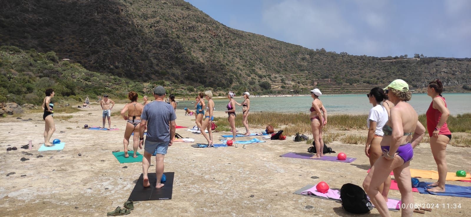 Persone che fanno esercizi su una spiaggia con un lago e colline sullo sfondo. Giornata di sole.