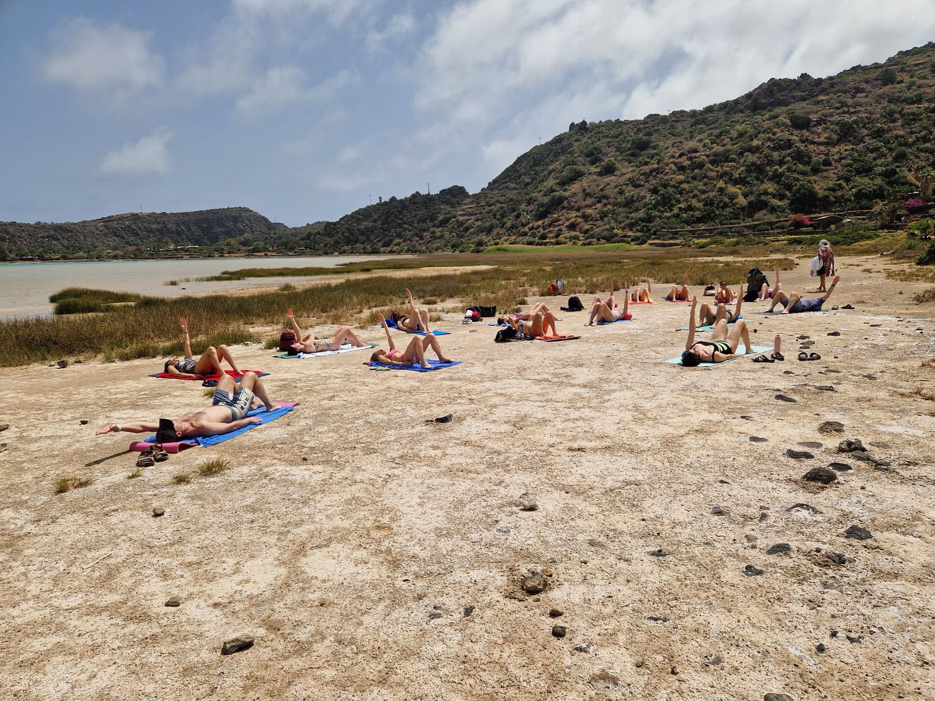 Persone che fanno yoga su una spiaggia sabbiosa vicino a un lago, con uno sfondo collinare sotto un cielo luminoso.