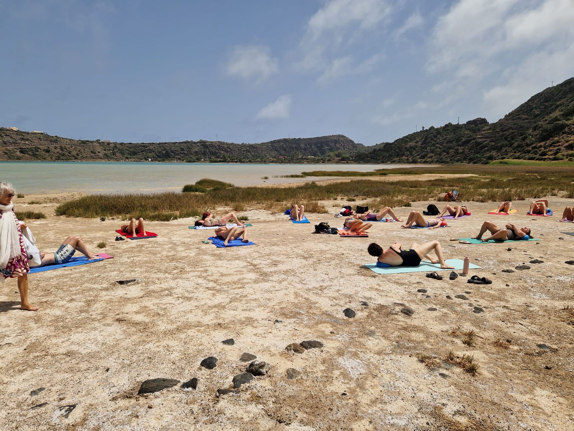 Gruppo che fa yoga su una spiaggia in riva a un lago, con le montagne sullo sfondo. Cielo luminoso e giornata di sole.