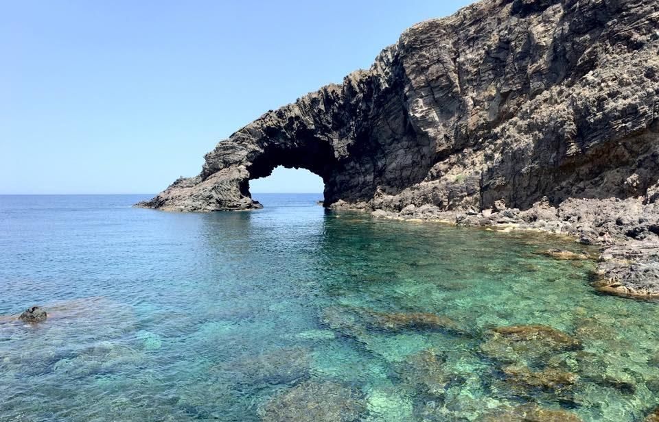 Arco di roccia costiera che si affaccia su acque turchesi e cristalline, con una costa rocciosa e un cielo azzurro.