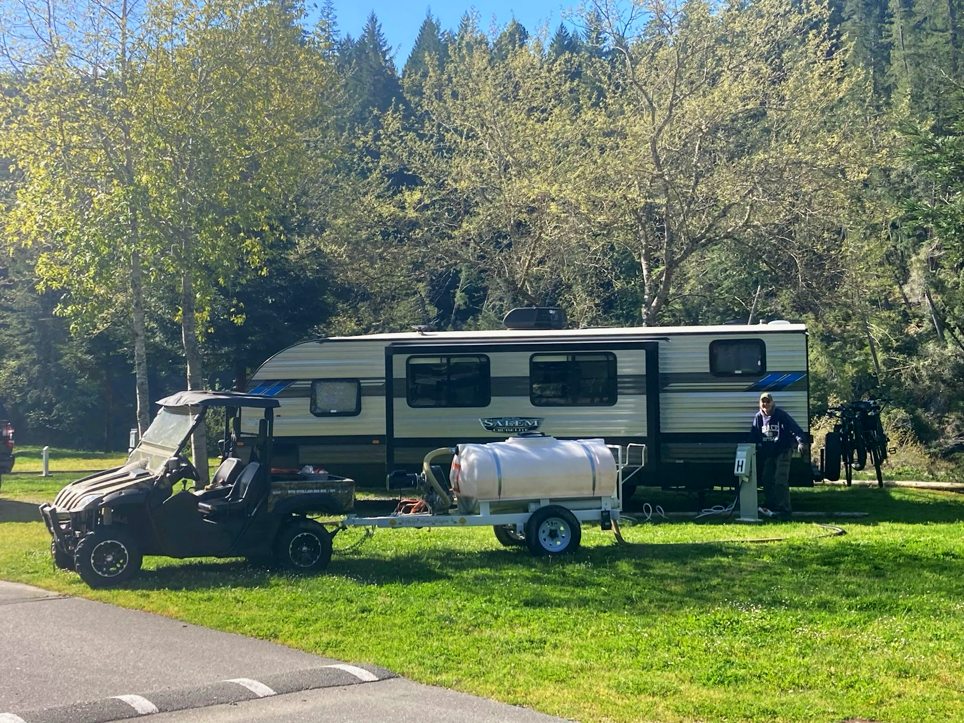 A large beige recreational vehicle parked on a green lawn by a river, shaded by trees under a blue sky.