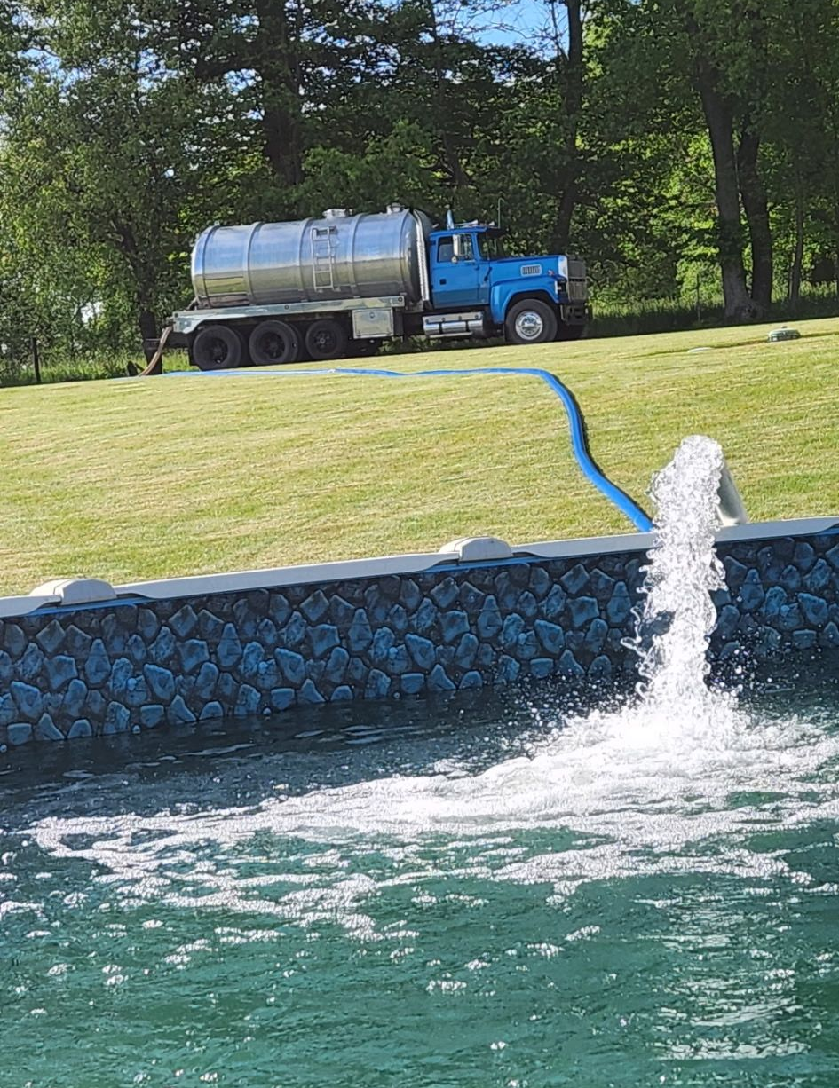 Blue septic truck filling a swimming pool with water via a blue hose on a grassy hill.