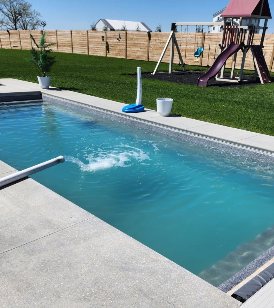A rectangular in-ground pool with clear water, surrounded by concrete and grass. A playset and fence are in the background.