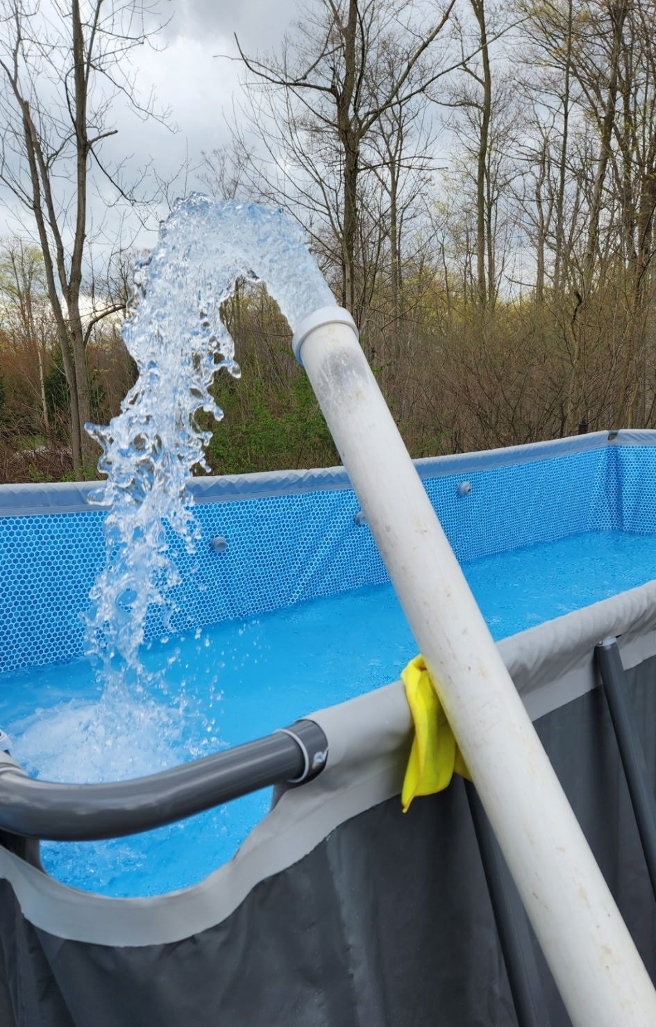 Water gushing from a white pipe into a blue-lined above-ground pool; a yellow cloth is clipped to the pool's side.