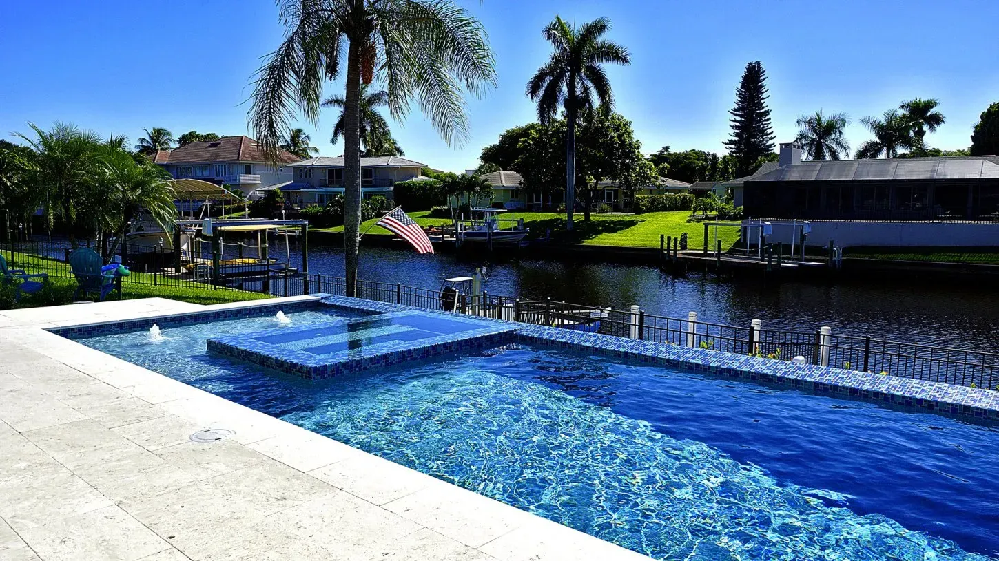 Swimming pool overlooking a canal, with blue water and palm trees under a sunny sky.