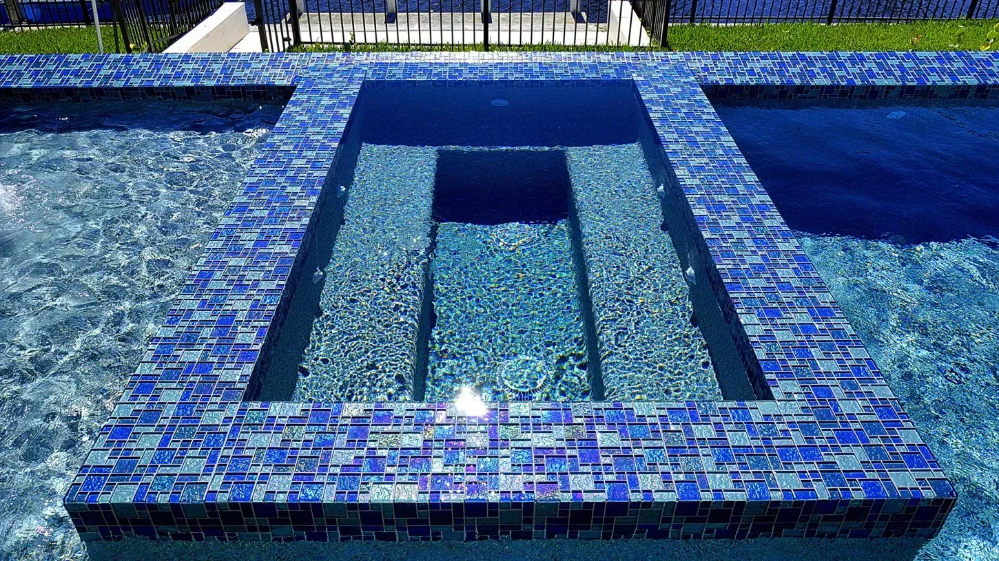 A blue mosaic-tiled pool with steps and a rectangular in-pool seating area; outdoor setting.