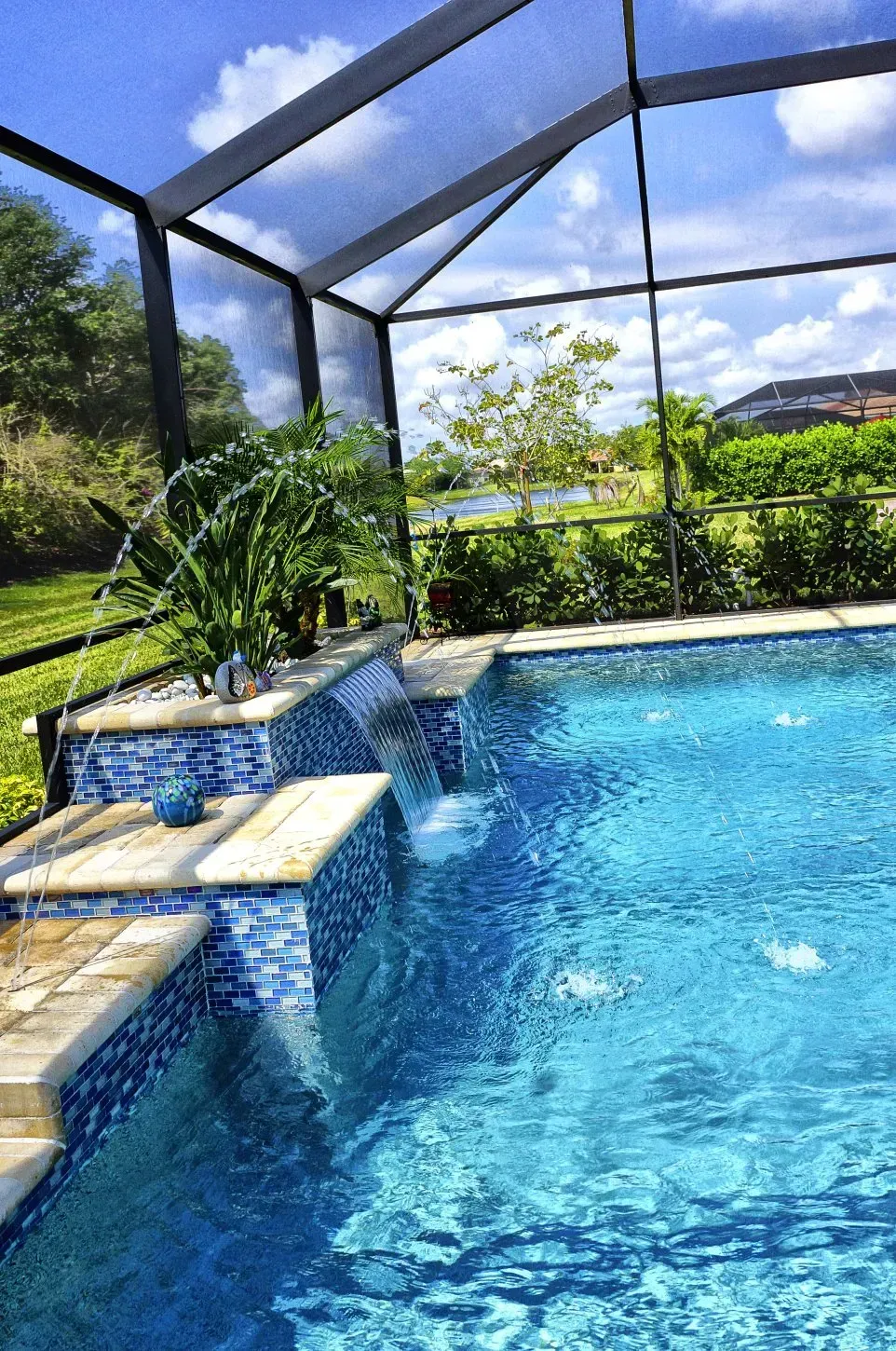 Pool with blue tiled waterfall, screened enclosure, and green plants. Sunny day.