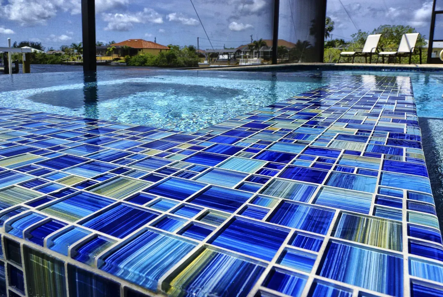 Close-up of a tiled pool border with varying shades of blue and green, overlooking water and distant trees.