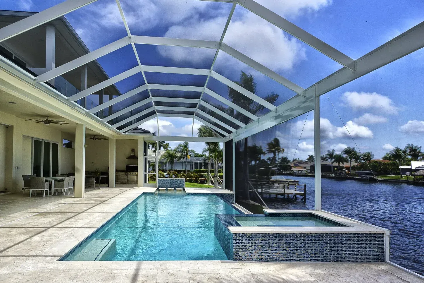 Pool and jacuzzi on a waterfront patio, under a screened enclosure, on a sunny day.