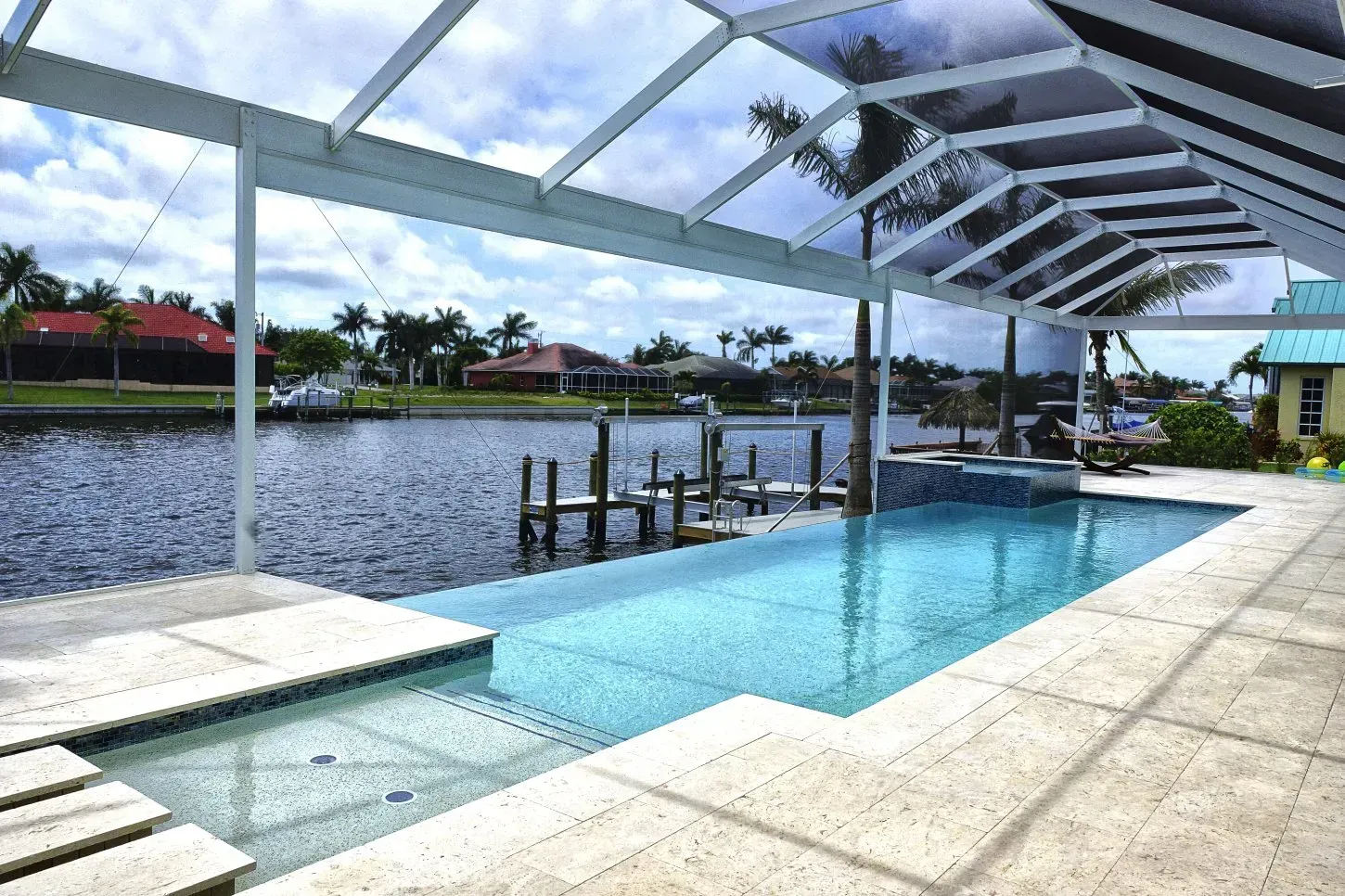 Poolside view of a canal with a pool, spa, and screened-in enclosure. Blue water, blue sky, and palm trees.