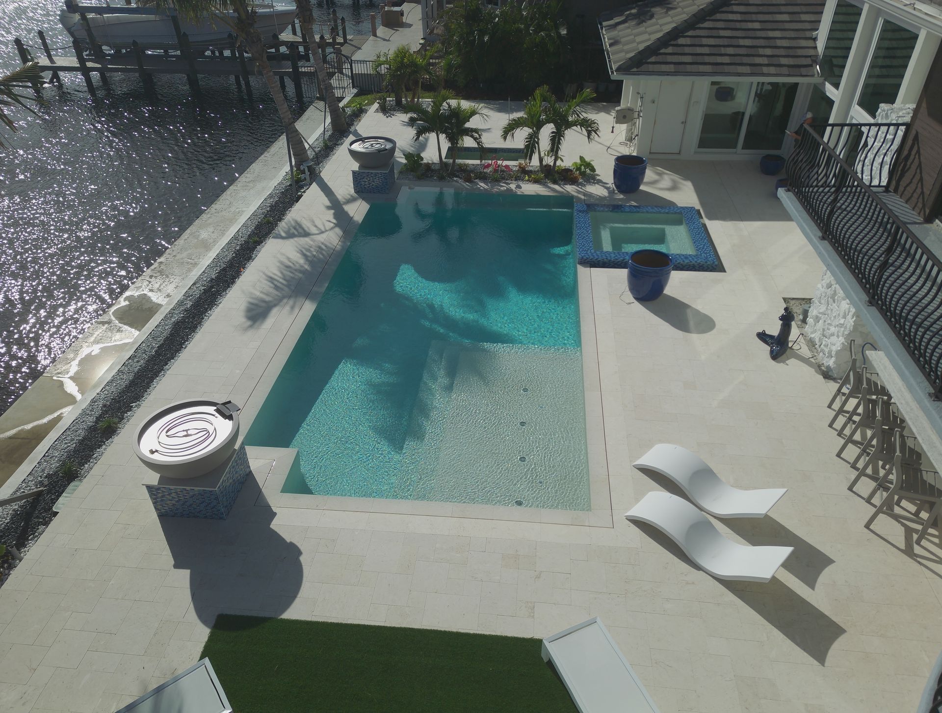 High-angle view of a pool and patio.  Pool is surrounded by white concrete, lounge chairs, and landscaping.  Adjacent to water.