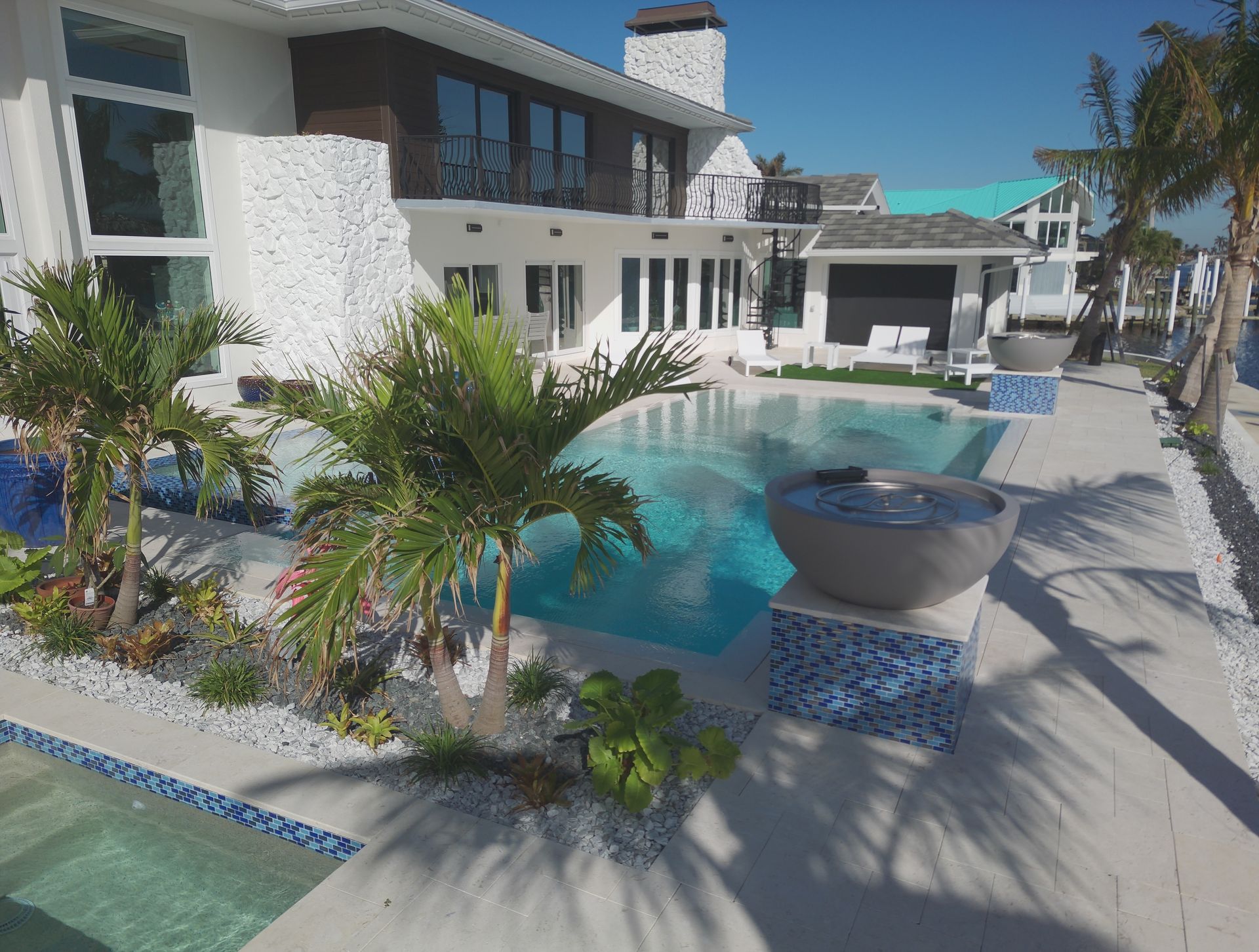 Luxury home with a pool, palm trees, and water view. White and blue accents in a sunny setting.
