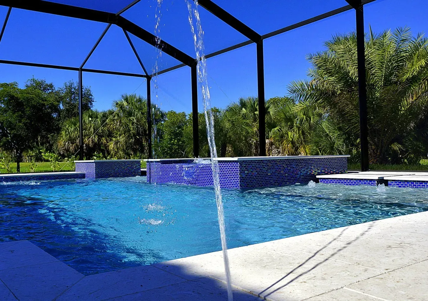 Swimming pool with blue tiles, water feature, blue sky, and screen enclosure.