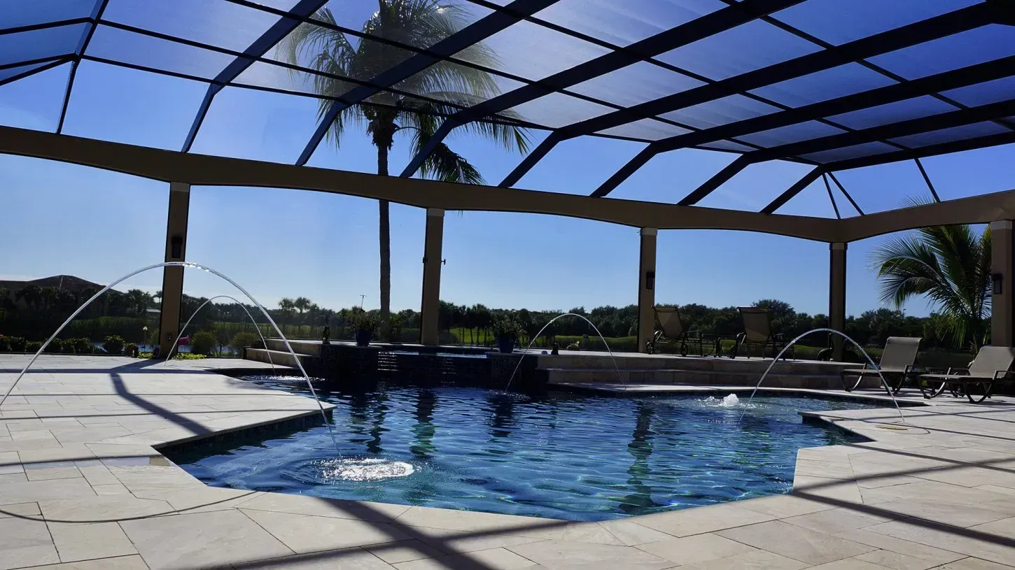 A swimming pool with a screened enclosure under a sunny sky. Water fountains and outdoor seating are visible.