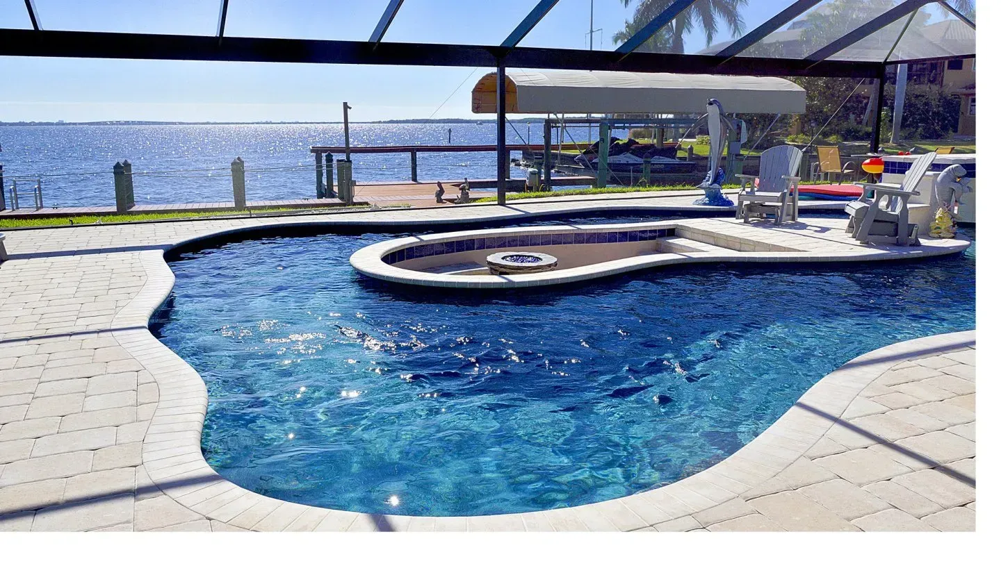 A curved pool with blue water overlooks a waterway; chairs sit on a raised deck area.
