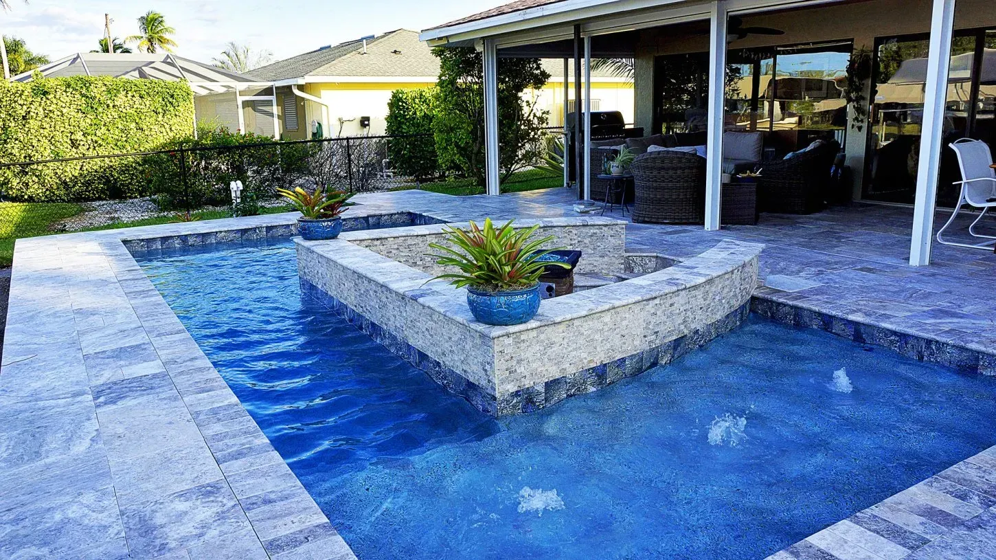 A luxury backyard pool with a raised spa and fire pit, with a house and greenery in the background.