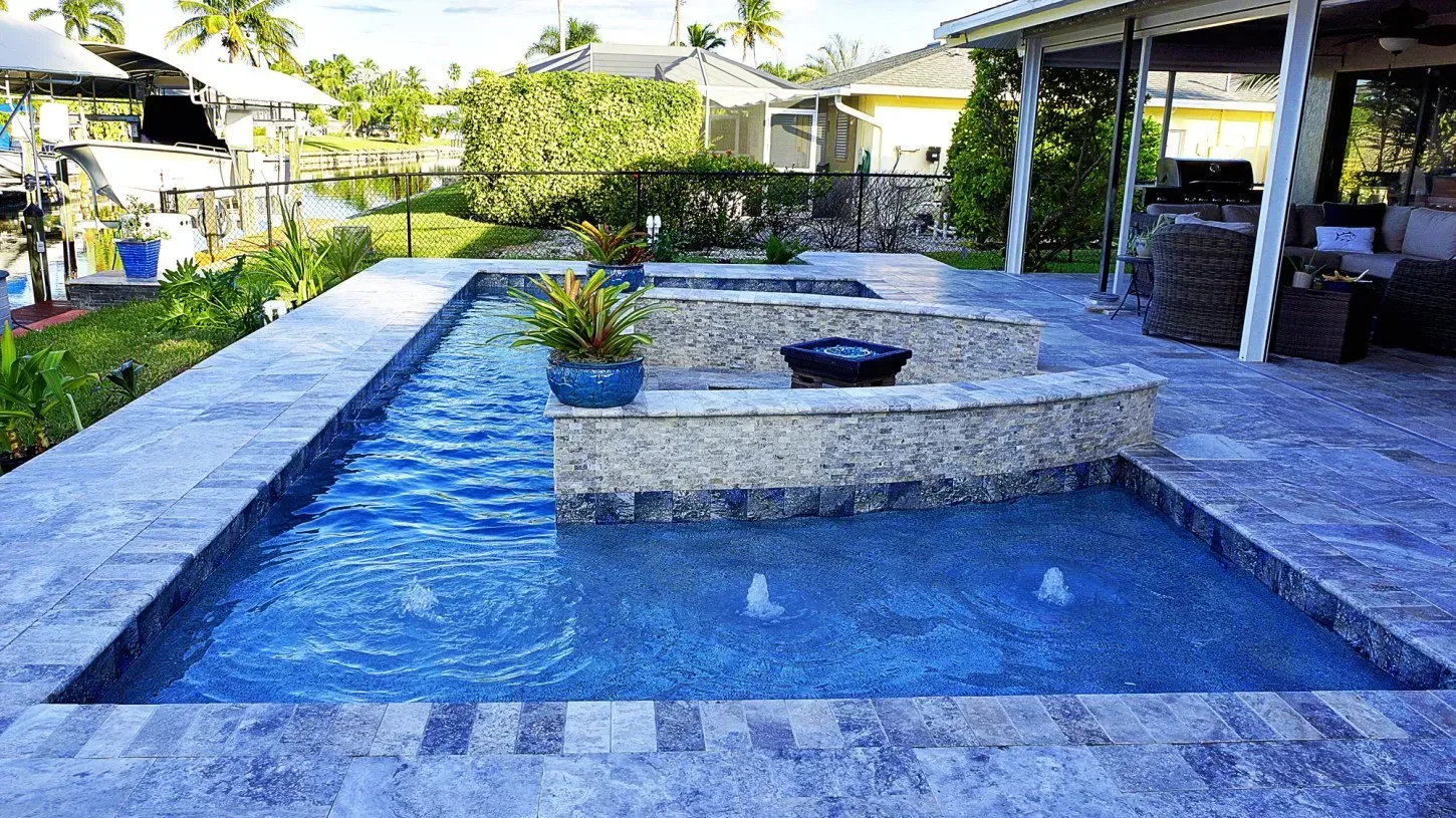A rectangular pool with water fountains and a stone ledge, in a backyard with a canal view.