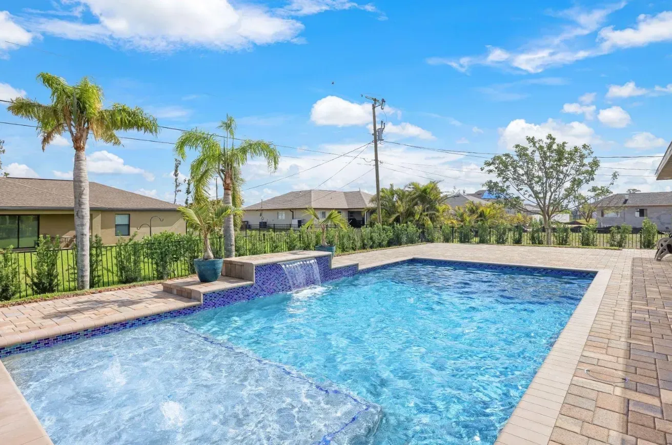 Backyard pool with a waterfall feature, palm trees, and a sunny sky.