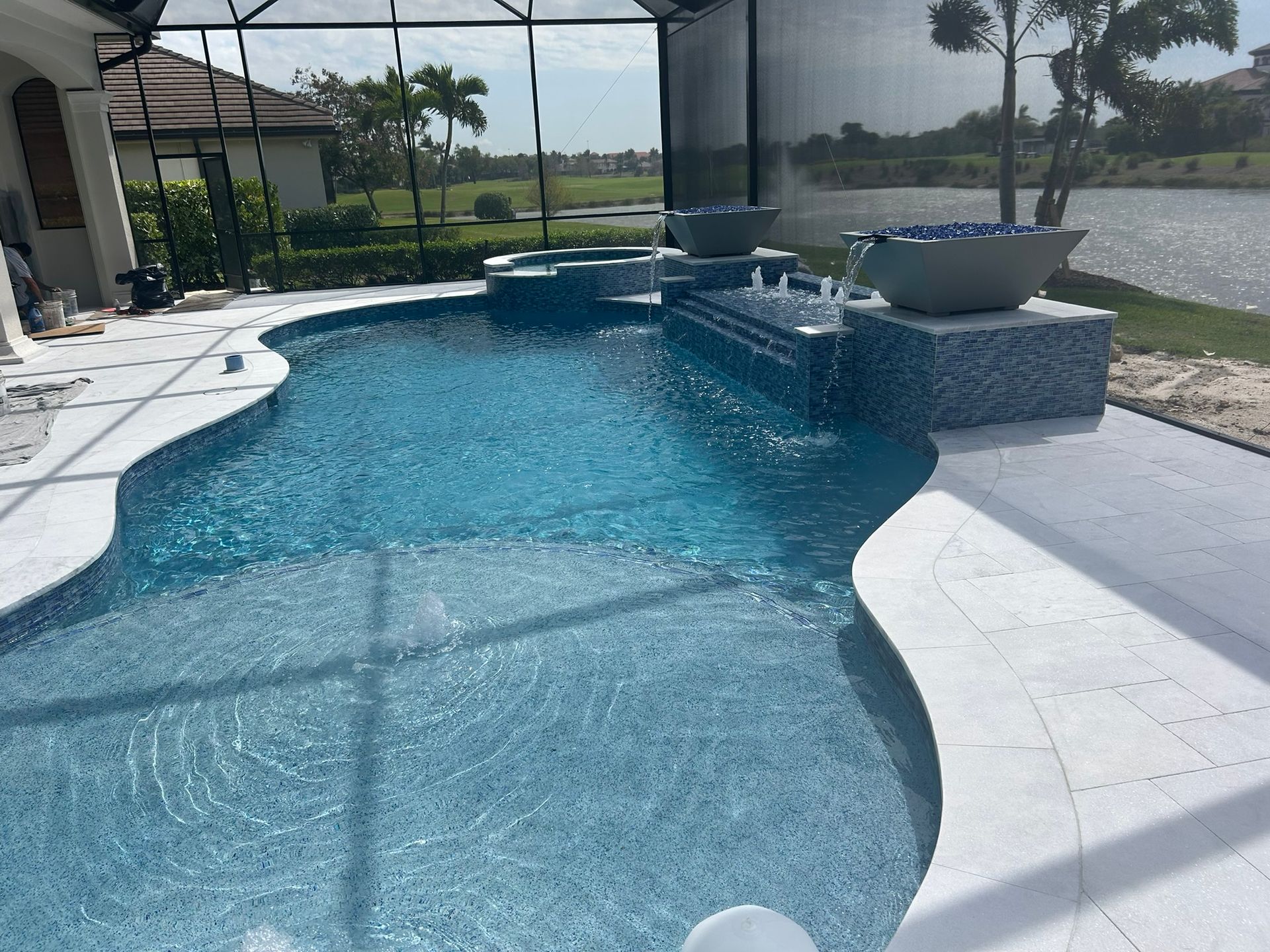 Swimming pool with blue tiles, water features, and light-colored patio, near a lake.