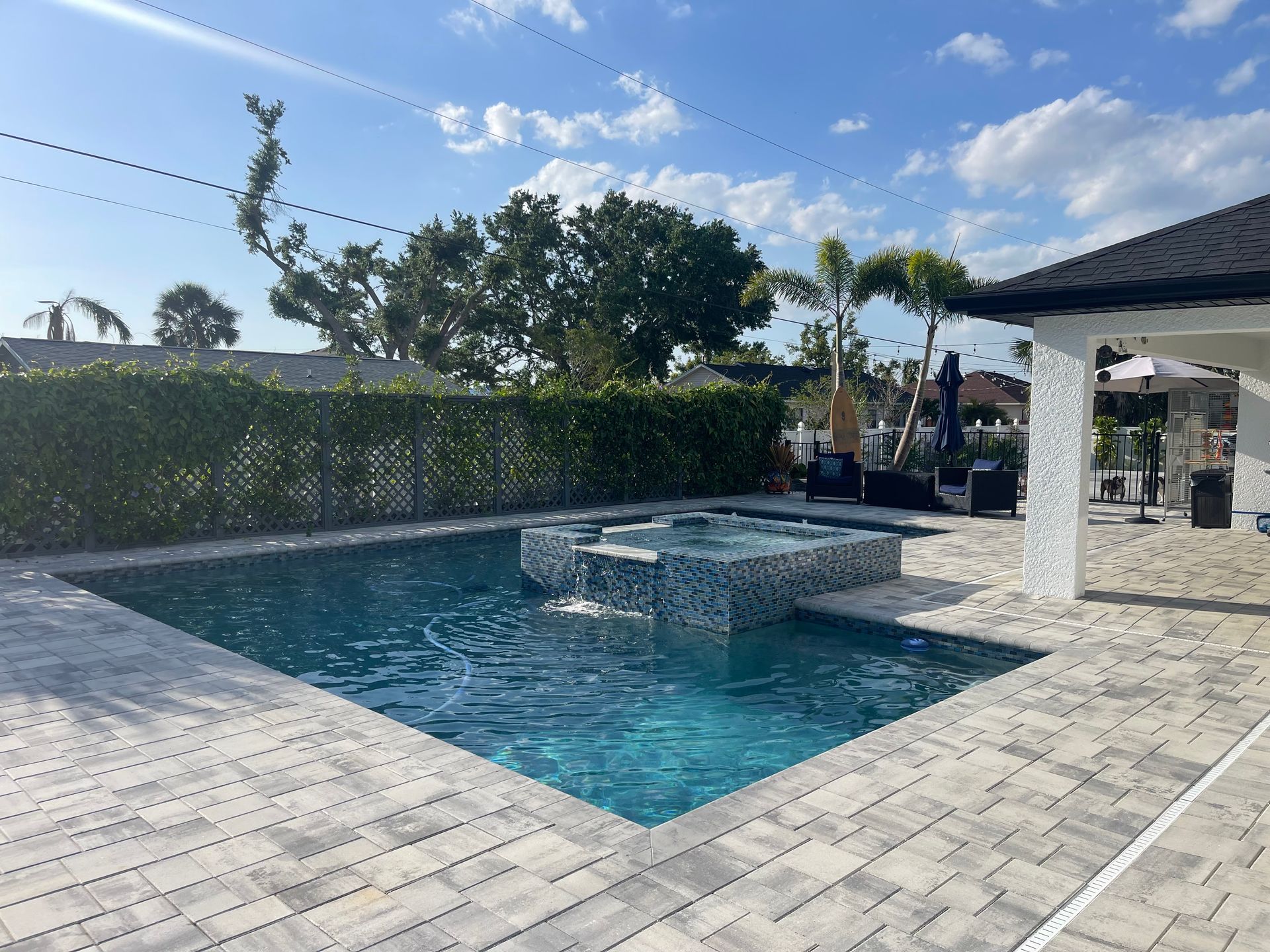 A pool with a spa, patio, and green hedge under a blue sky.