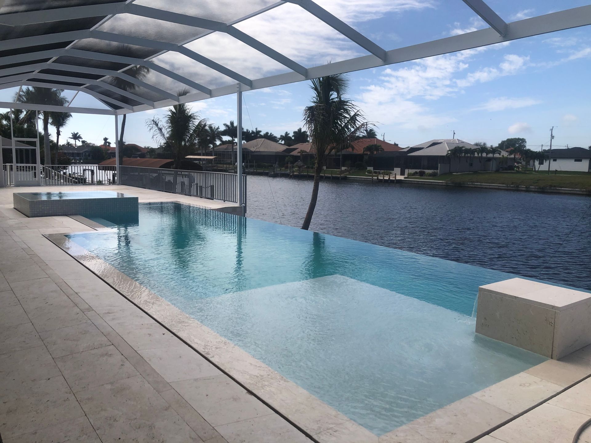 Infinity pool overlooking a canal, with a white pergola, blue water, and palm trees.
