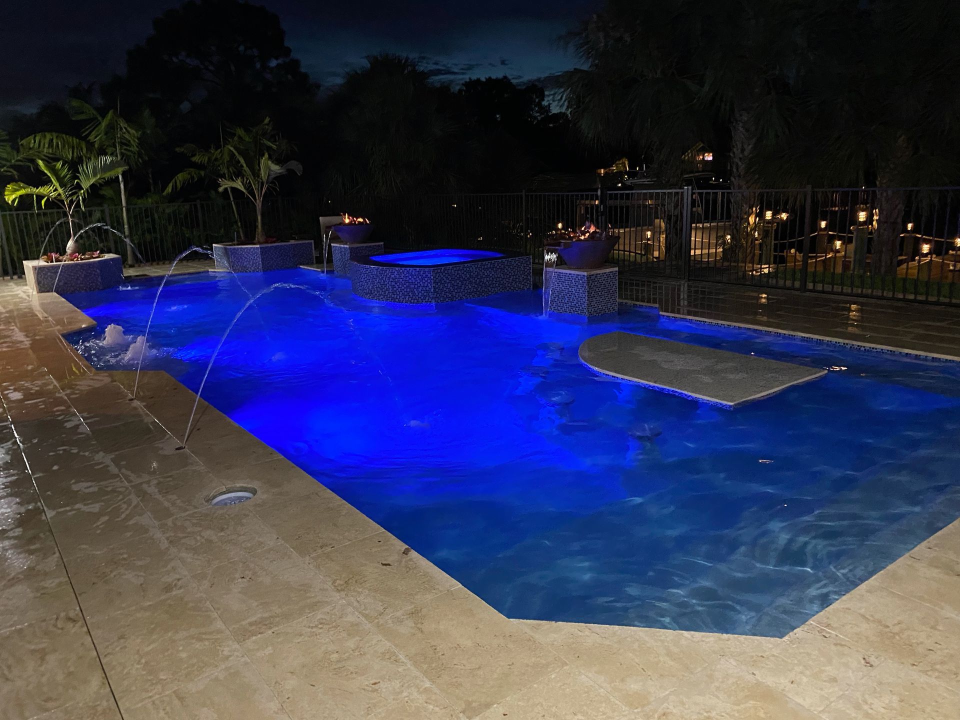 Nighttime view of a pool illuminated in blue with fountains, surrounded by a patio and trees.